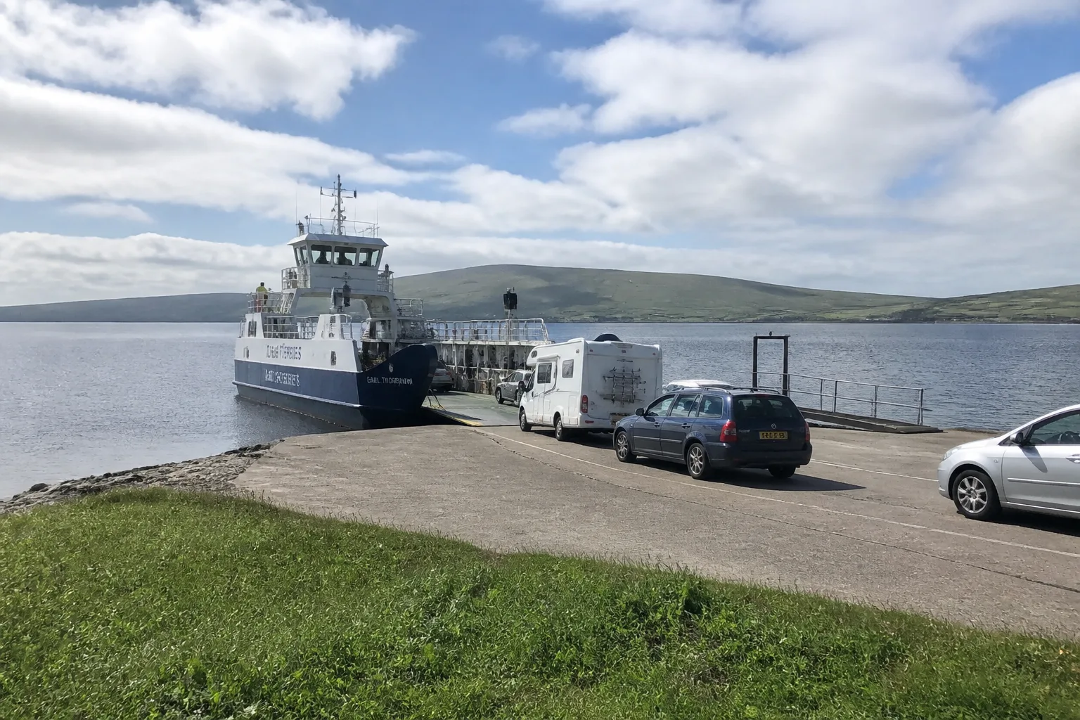 Small Orkney Ferries roll-on roll-off vessel arriving at Tingwall slipway on Mainland Orkney, with bow ramp lowering and a queue of cars waiting to board, low blue-grey hills of Rousay visible in the background