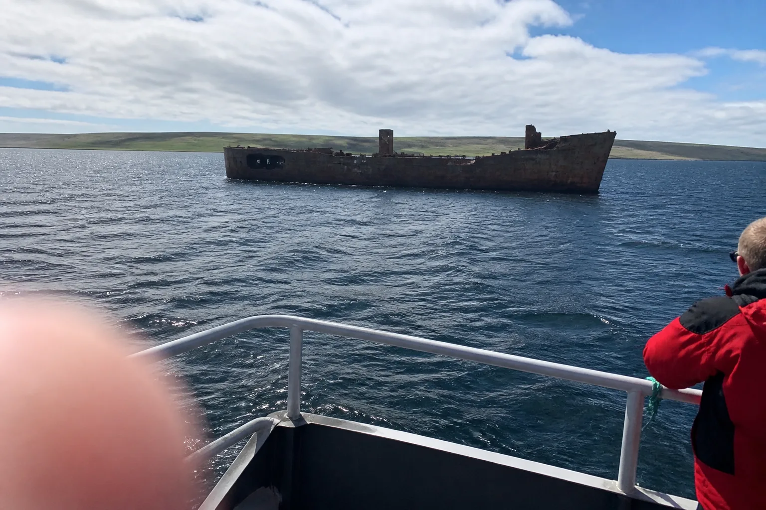 Rusted hull of an old wartime blockship rising out of the choppy steel-grey water of Scapa Flow, photographed from a small wildlife boat with low green coastline in the background