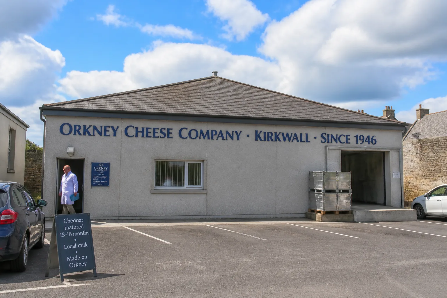 A mid-range photo from the road in front of the Orkney Cheese Company creamery on Crowness Road in Kirkwall — a modern but plain industrial building with grey rendered walls and a low-pitched grey-tile roof with a large painted sign in dark navy lettering reading ORKNEY CHEESE COMPANY KIRKWALL SINCE 1946, a small loading bay with stainless steel milk-handling crates beside it, a tarmac car park in the foreground with a wooden A-board on the pavement reading Cheddar matured 15-18 months Local milk Made on Orkney, under a pale-blue summer sky with broken cloud