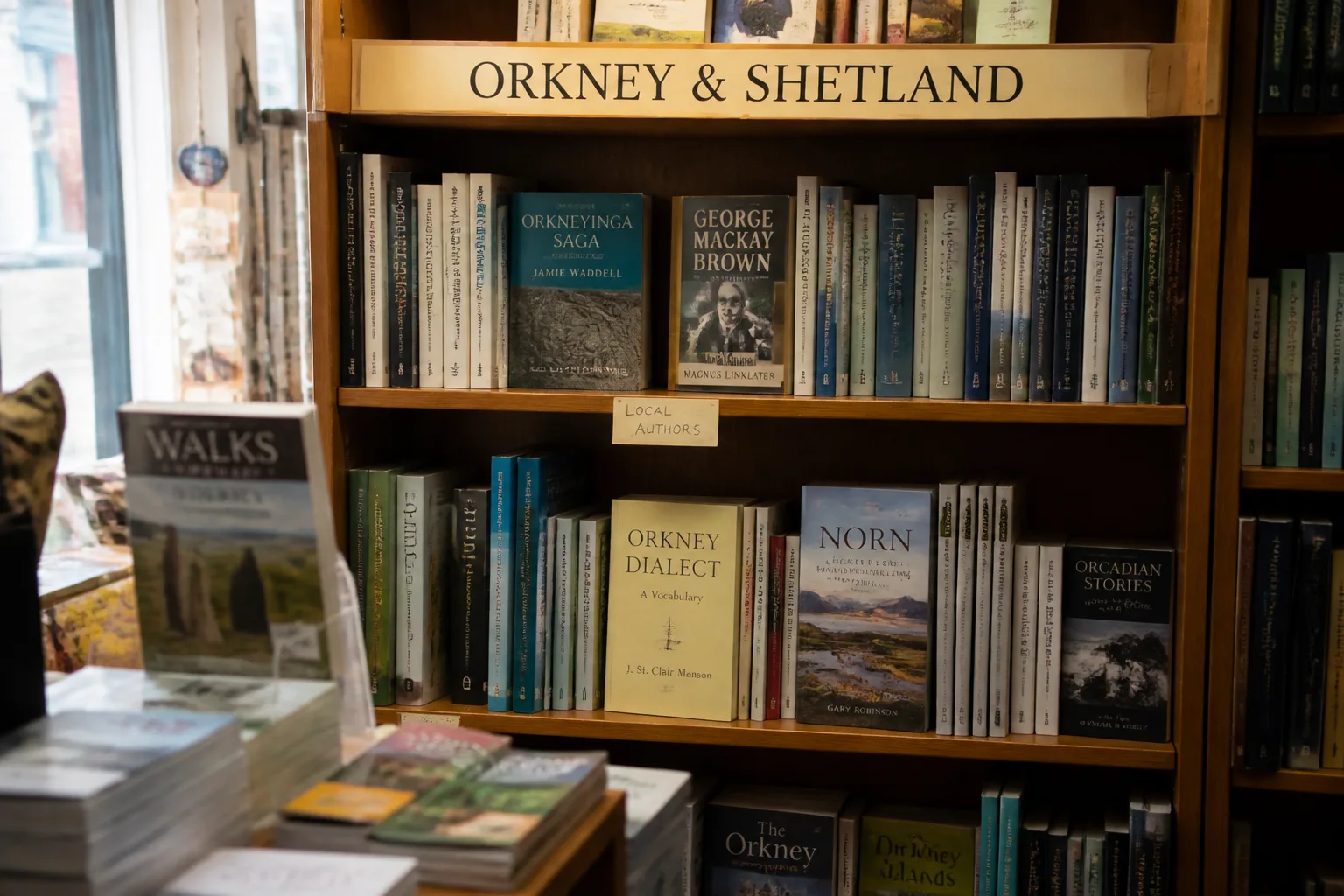 A wooden bookshelf inside a small independent bookshop in Kirkwall, Orkney, showing an Orkney and Shetland regional-interest section with paperback spines including Orkneyinga Saga, George Mackay Brown, Orkney Dialect, Norn and Orcadian Stories, with a hand-written LOCAL AUTHORS cardboard tab between shelves and warm natural side-light from a shop window