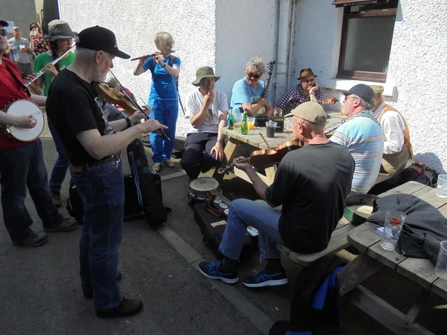 Musicians with fiddles, flutes and a banjo playing an informal outdoor session on picnic benches outside the Ferry Inn in Stromness during the Orkney Folk Festival