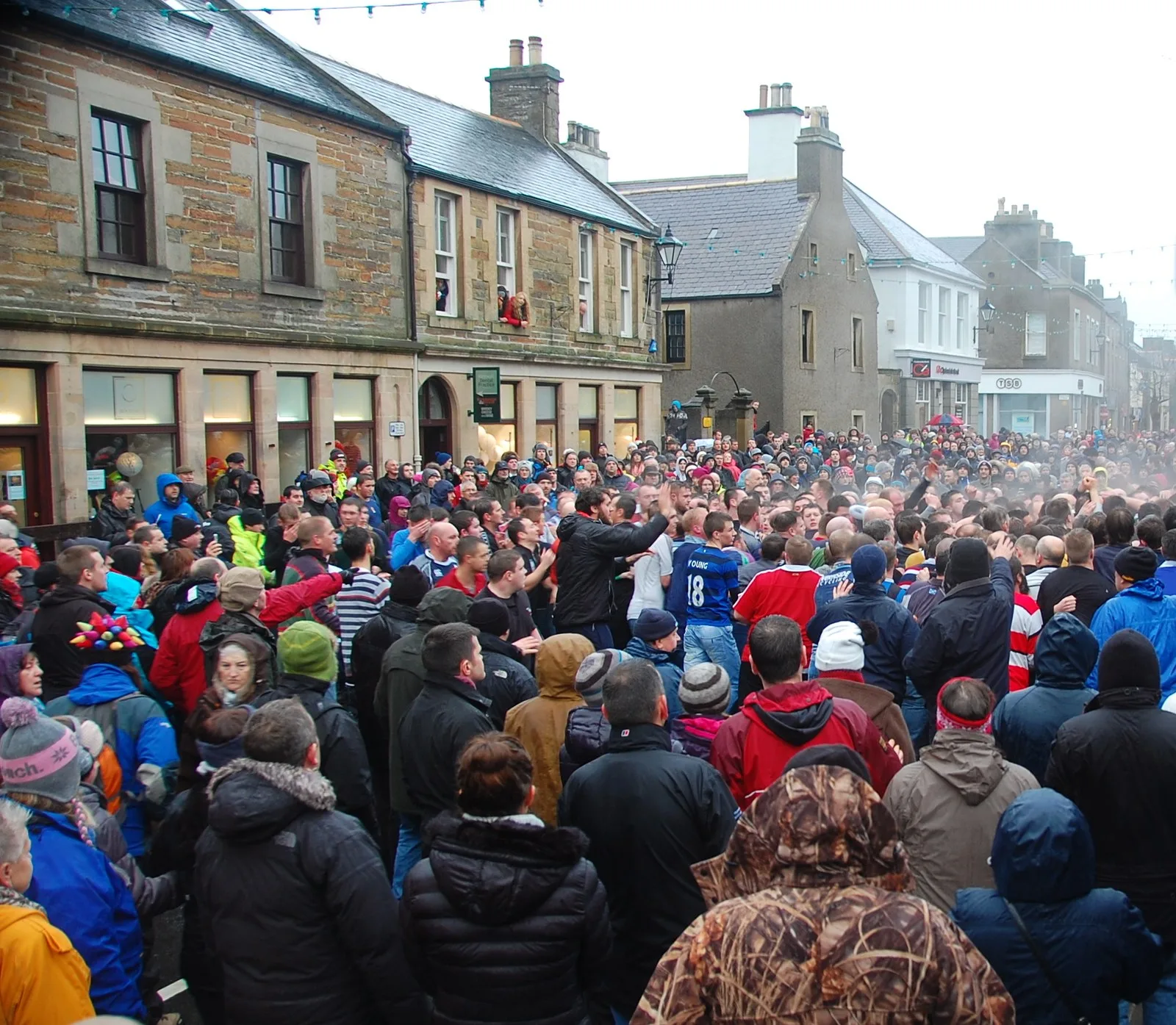 A dense crowd packed into Broad Street, Kirkwall, for the New Year's Day Men's Ba', with boarded shopfronts and spectators watching from upstairs windows