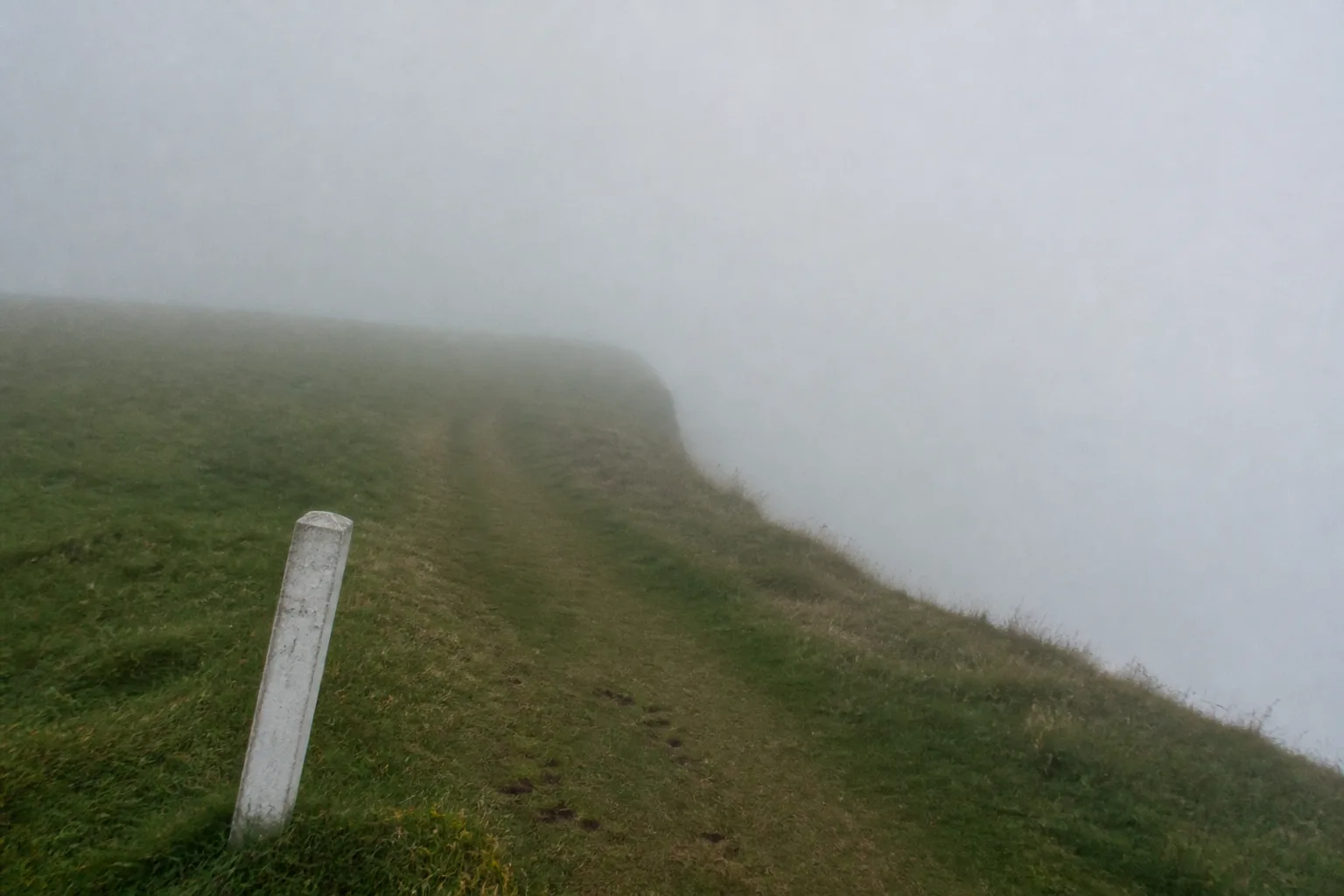 An empty grass coastal path along the cliff edge at Marwick Head, Orkney, photographed in thick afternoon mist with the path stretching into white blank fog and a single white-painted wooden marker post leaning slightly in the foreground — the bleak oppressive mood typical of Nuckelavee territory in local folklore