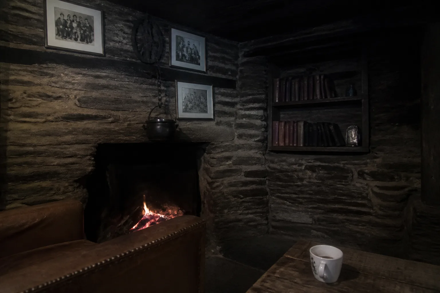 A small peat fire burning in a stone fireplace inside an old Orkney longhouse-style cottage at night with a warm orange glow from the embers, a black iron cooking pot on a chain above the fire, framed black-and-white photographs of Orkney crofters on the stone wall, and a corner of a worn brown leather armchair in the foreground