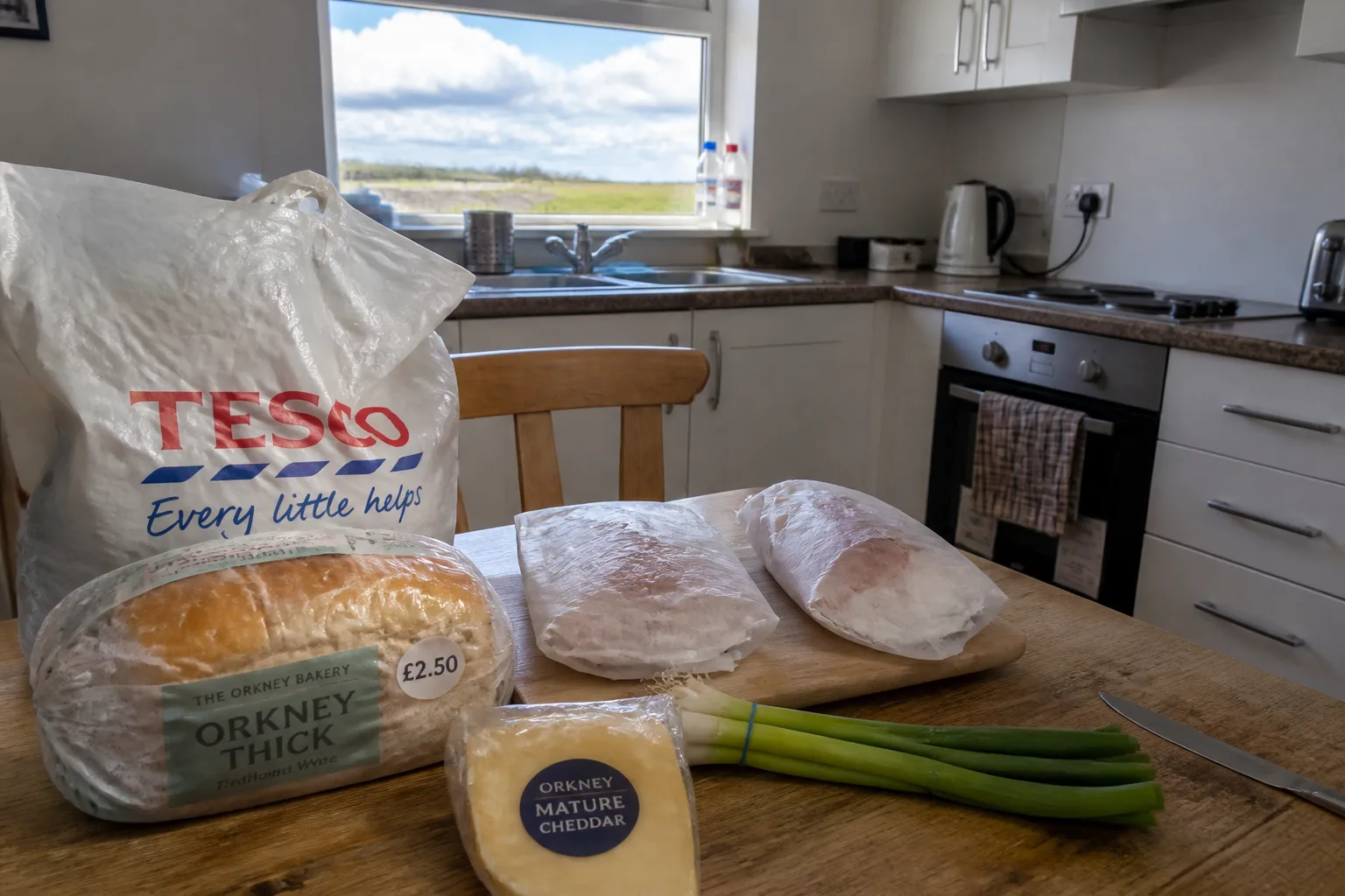 Self-catering cottage kitchen in Orkney mid-meal-prep with a Tesco carrier bag on the wooden table, a £2.50 Orkney bread loaf, a wedge of Orkney mature cheddar, fresh haddock fillets on a board and a bunch of spring onions, with a window above the sink looking onto green farmland