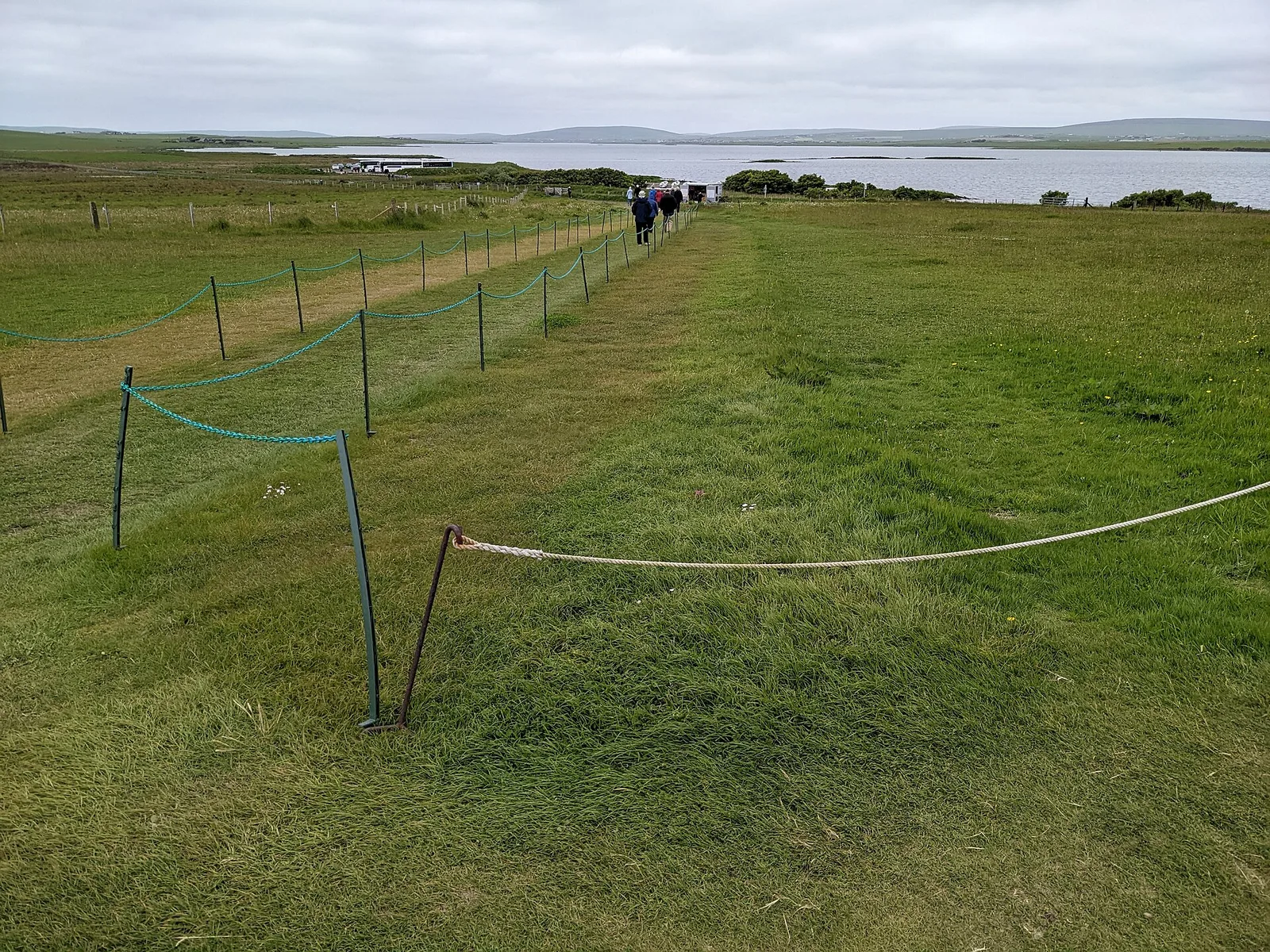 Roped-off grass paths at the Ring of Brodgar in Orkney, with visitors walking towards the loch and the car park