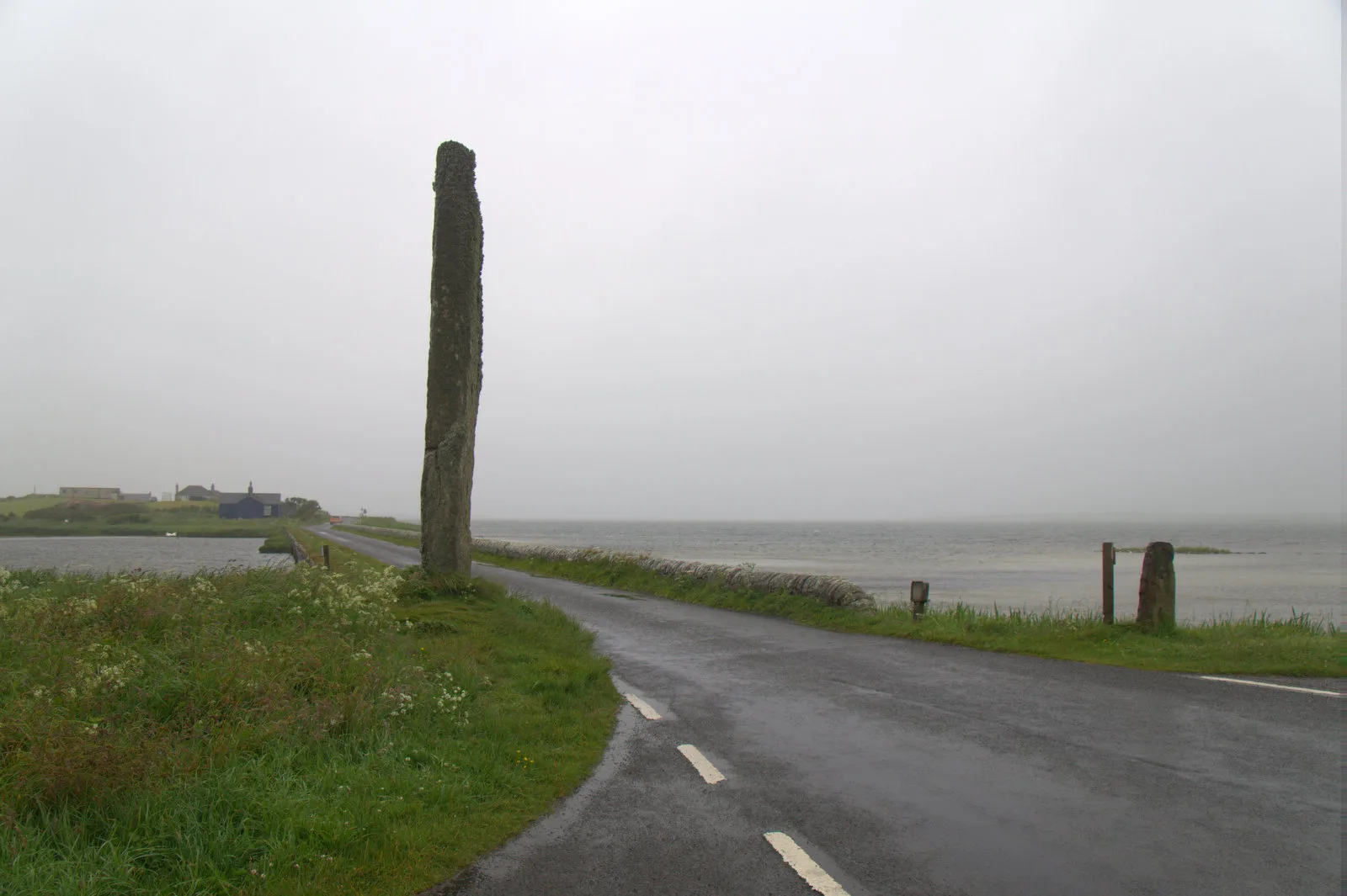The tall Watch Stone beside the road at the Bridge of Brodgar in Orkney, with loch water on the right on a wet, misty day