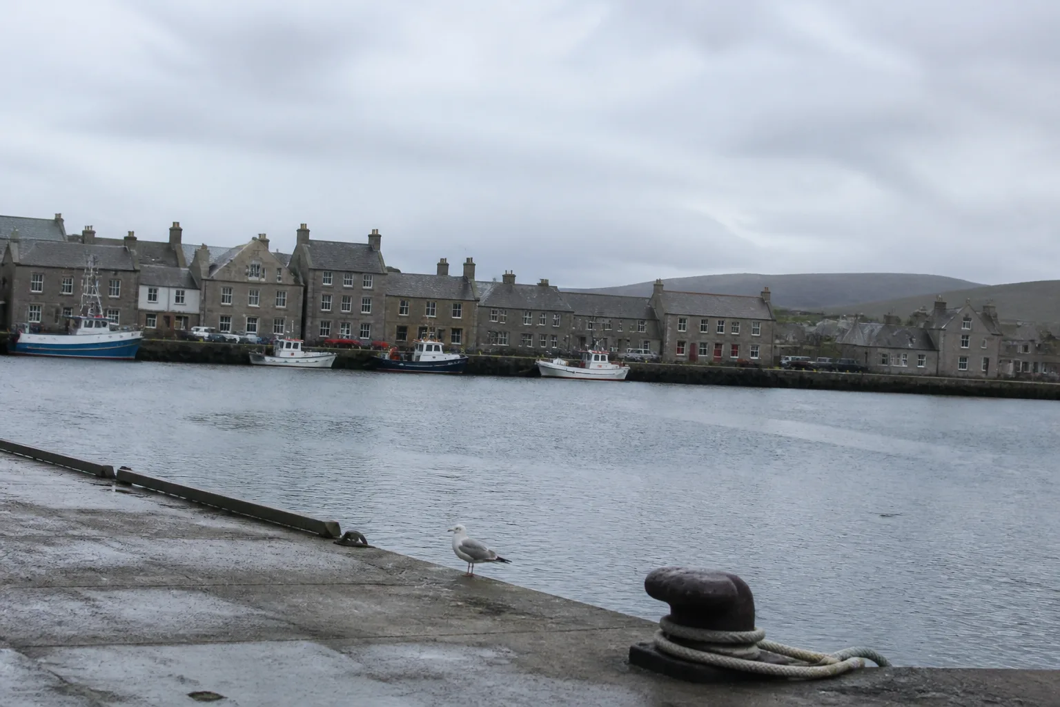 Small Northern Isles harbour with stone-fronted houses lining the waterfront, slate roofs, fishing boats moored at the pier and a wet stone bollard with coiled rope in the foreground — could be either Lerwick in Shetland or Stromness in Orkney