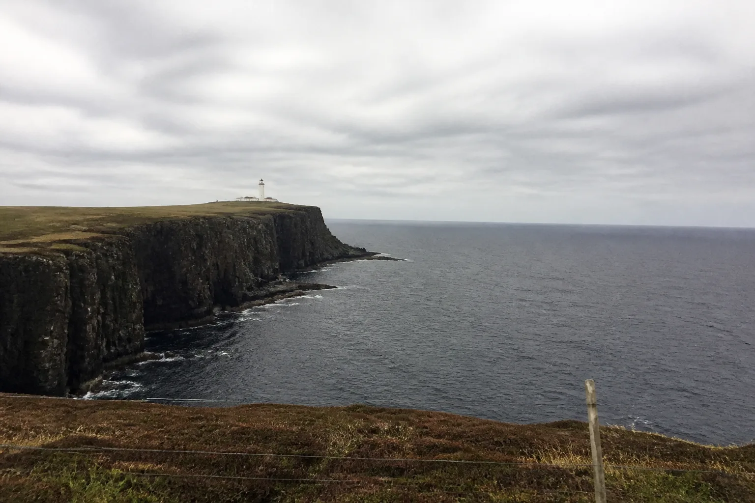 The cliff path at Sumburgh Head, the southernmost tip of mainland Shetland, with dramatic dark sandstone sea cliffs dropping straight to the steel-grey North Atlantic and the white Sumburgh Head lighthouse on the highest point under a grey-white sky
