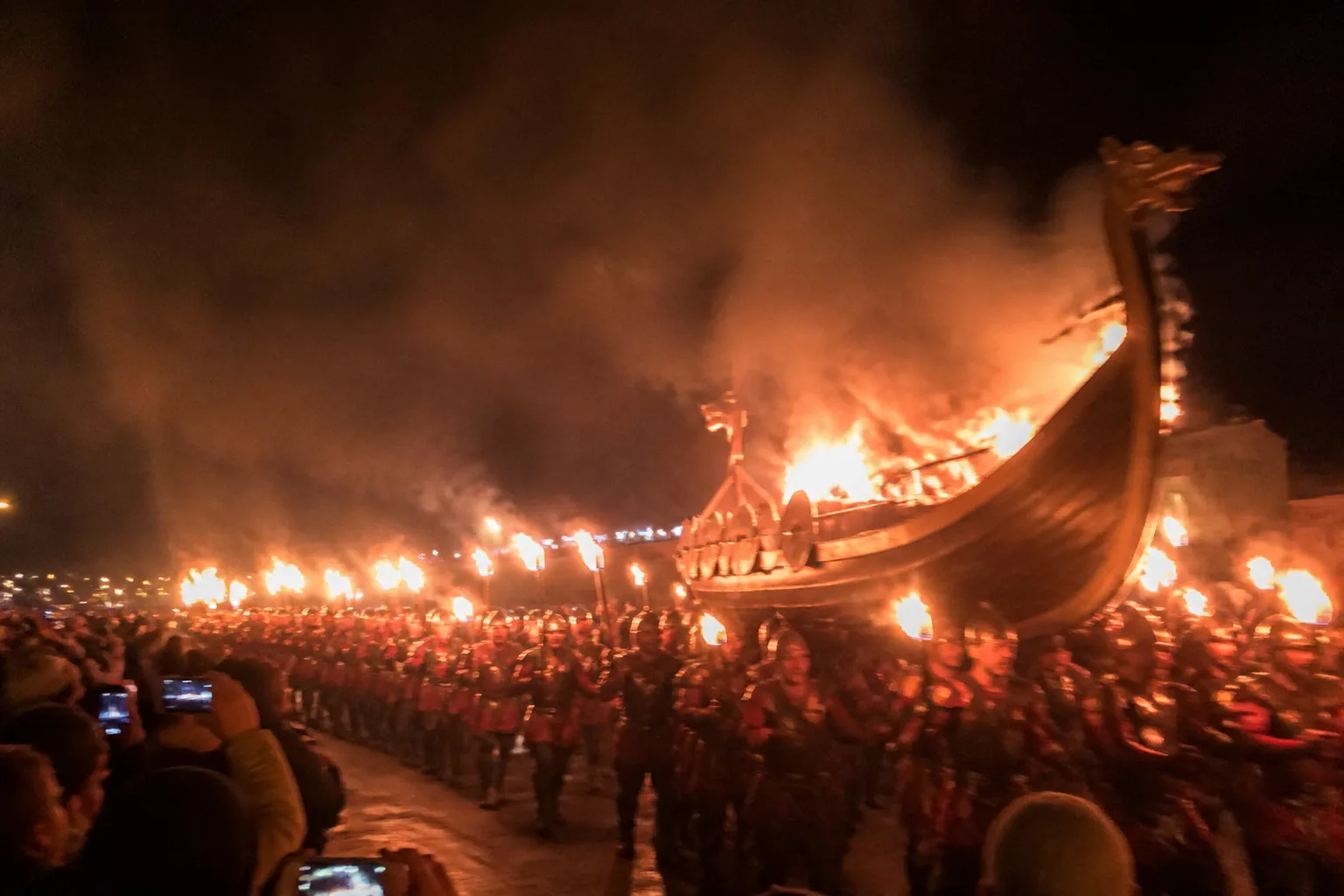 The Up Helly Aa fire festival in Lerwick, Shetland, on a January night with a long line of torch-bearing men in Viking costume walking alongside a wooden longship galley as it begins to burn — the festival climax — with a crowd of dark silhouettes and rising smoke