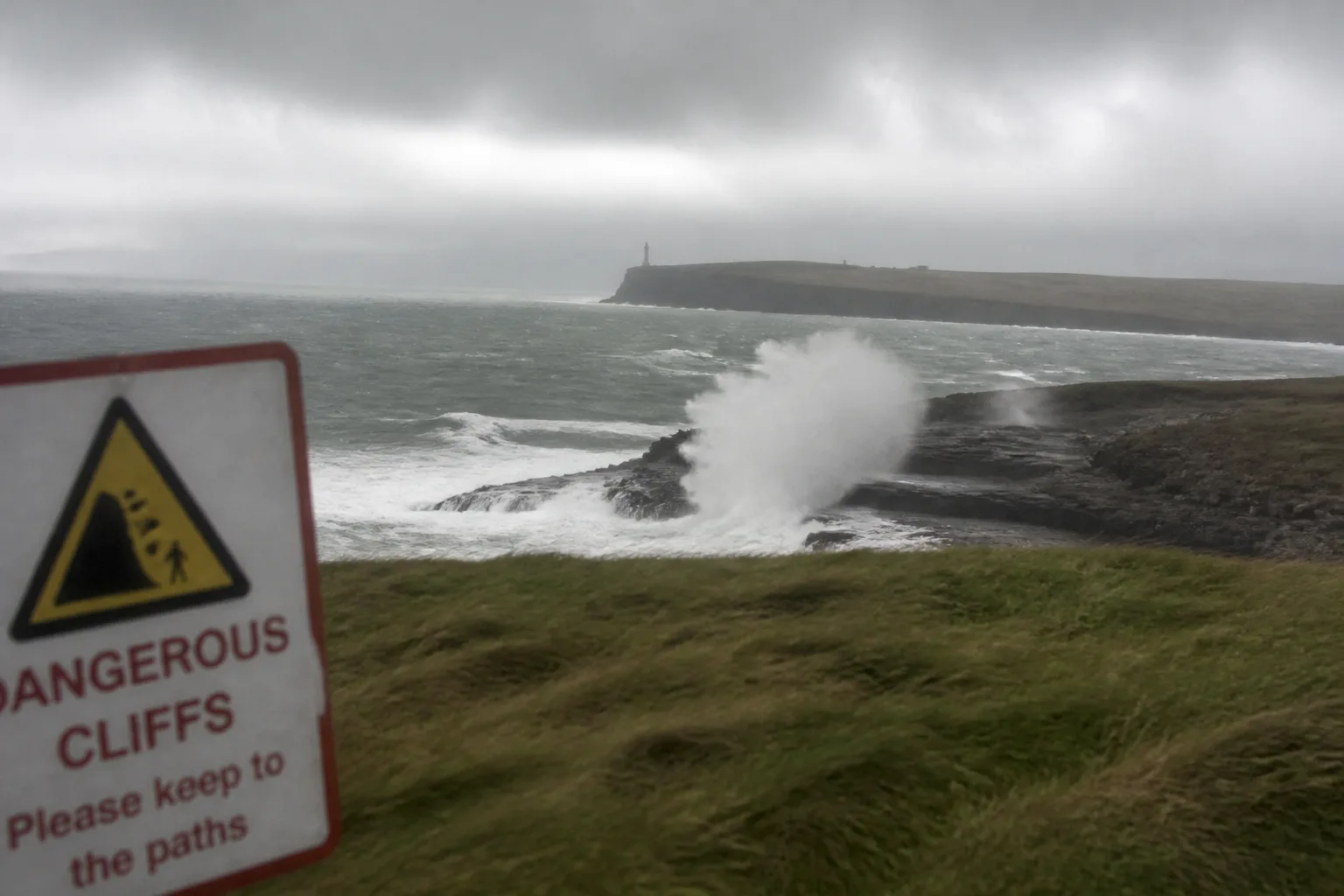 Huge white-capped Atlantic waves smashing into dark basalt rocks below Birsay Bay in Orkney during an October gale, throwing spray ten metres into the air, with the Brough of Birsay lighthouse just visible in low cloud