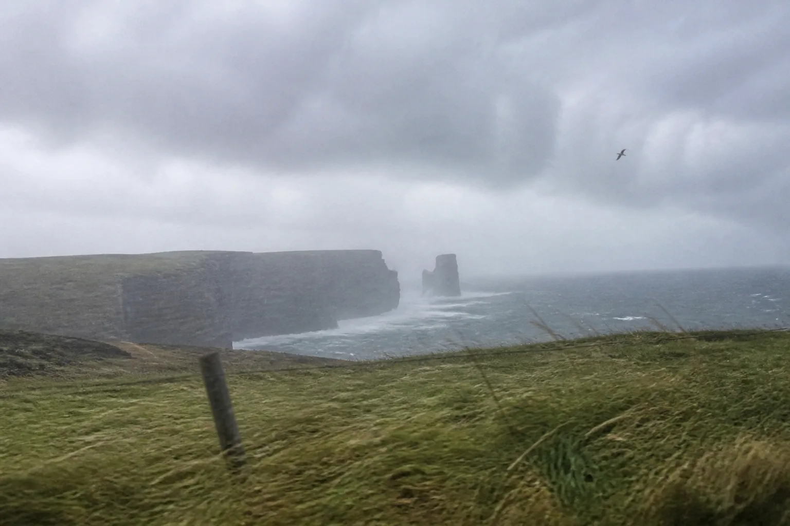 Stormy autumn day at Yesnaby cliffs, West Mainland of Orkney — horizontal rain blowing across the dark sandstone sea cliffs with the Yesnaby Castle sea stack just visible through the rain haze, a fence post leaning at an angle in the foreground