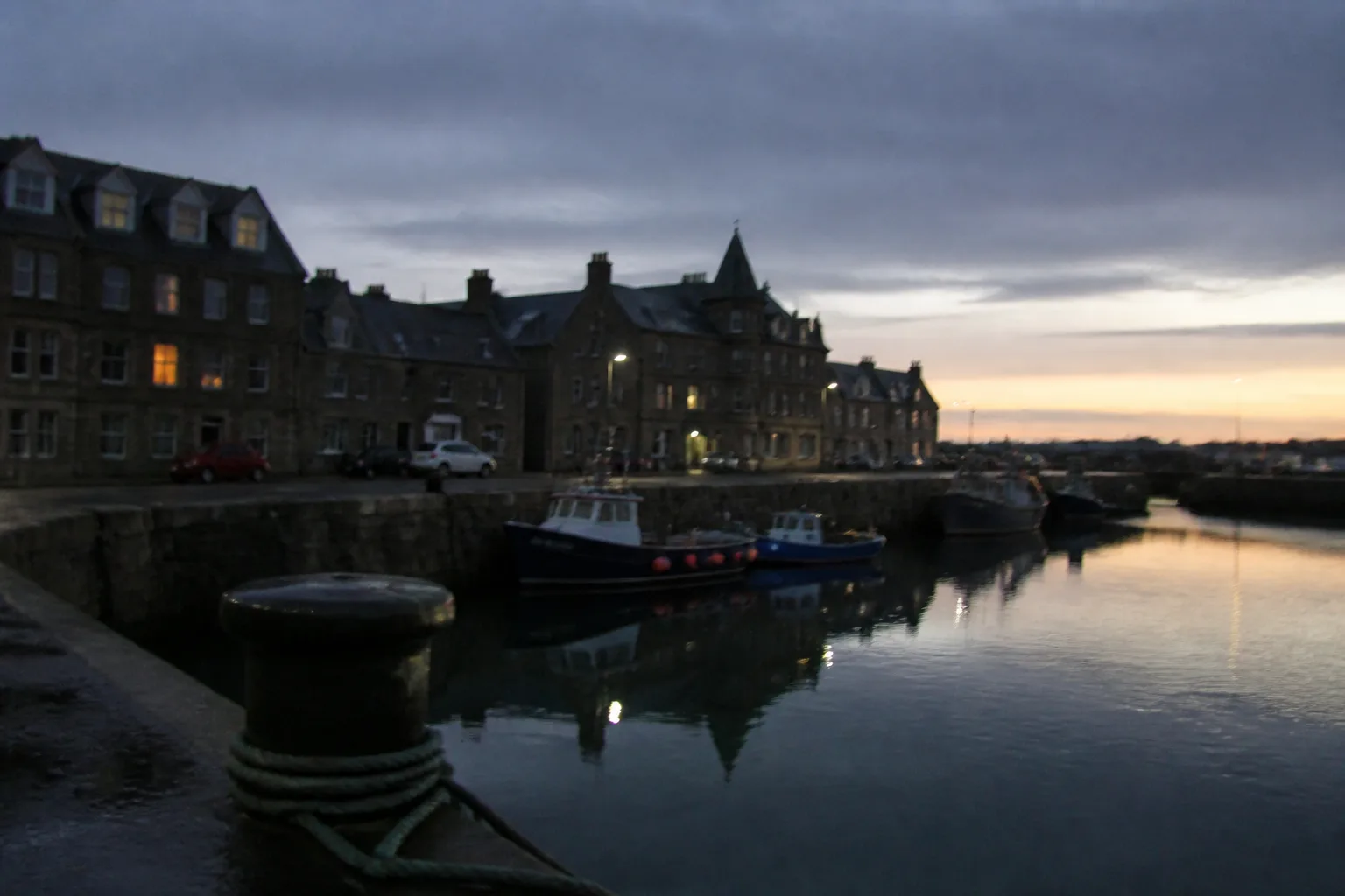 Stromness harbour at 11pm in mid-June during the Simmer Dim — sky still dimly luminous pale blue-grey with a soft orange band low on the horizon, fishing boats moored at the stone pier reflected in calm dark water