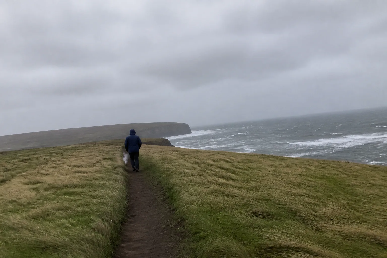 A single figure in a navy waterproof jacket and tight hood walking head-down along the clifftop path at Marwick Head, Orkney, leaning forward into the wind with the surrounding grass flattened sideways