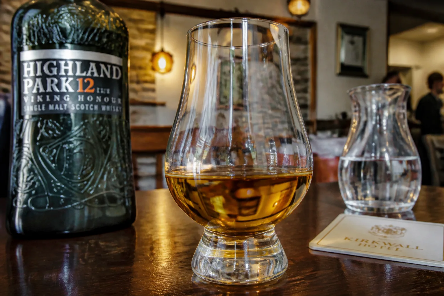 A close-up Canon DSLR photo of a Glencairn whisky glass holding a generous dram of amber-gold Highland Park 12-year-old single malt on the polished dark-wood bar of a Kirkwall hotel, with the dark green-and-gold Viking Honour bottle slightly out of focus behind, warm pendant-lamp light on exposed sandstone in the background