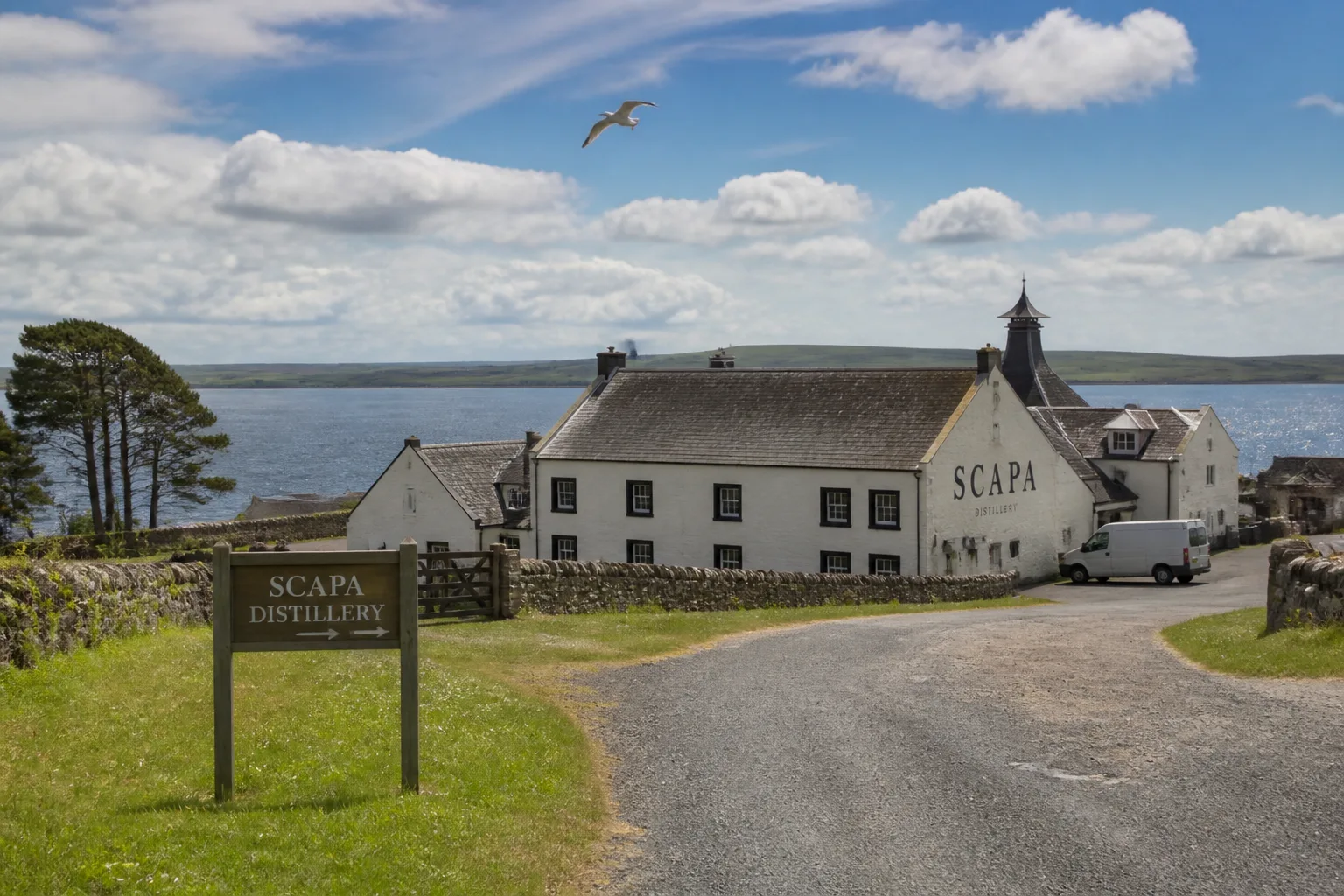 A wide Canon DSLR photo of Scapa Distillery at St Ola near Kirkwall, Orkney — the cream-painted main building with black-trimmed windows in the middle distance, the dark calm waters of Scapa Flow visible beyond, and the low green outline of the island of Flotta on the horizon under a sky of broken mid-June cloud with patches of bright blue
