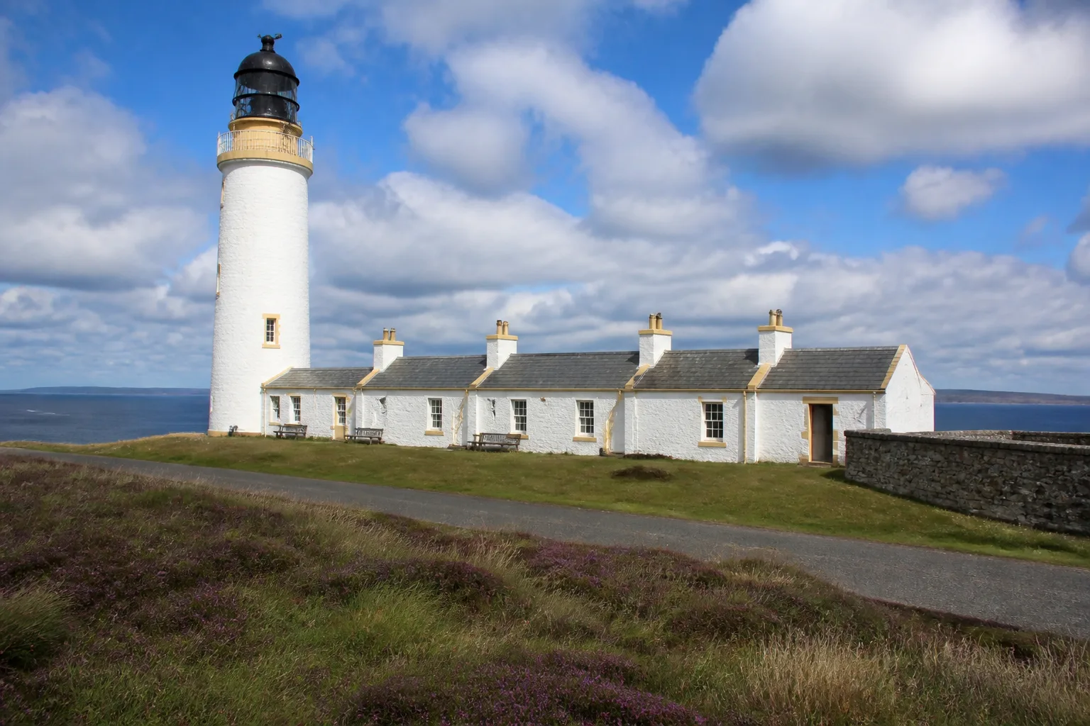 Cantick Head lighthouse and its row of single-storey whitewashed former keepers' cottages on the South Walls headland of Hoy, Orkney, with the Pentland Firth visible in the background under a partly-cloudy summer sky