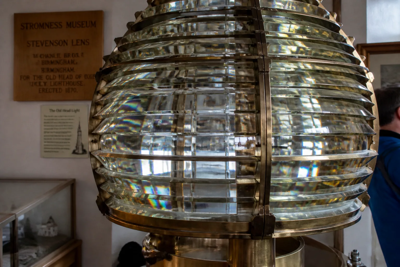 Close-up of an antique Stevenson-era Fresnel lens displayed inside Stromness Museum, Orkney, showing concentric brass-mounted glass prism rings stacked around a central optical core