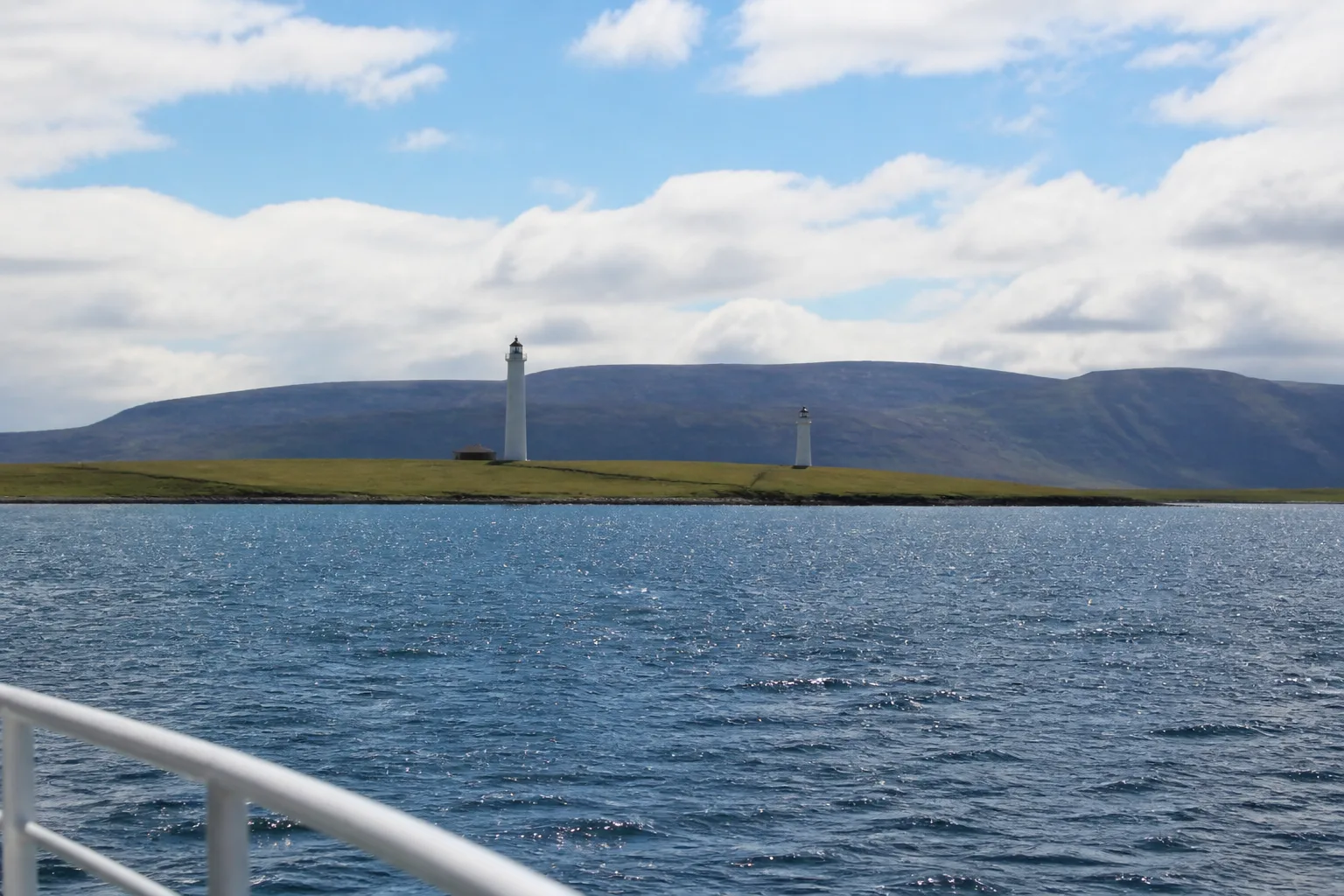 View from the passenger ferry deck looking across the blue waters of Hoy Sound toward Graemsay island in Orkney, showing the twin lighthouse towers of Hoy Sound High and Hoy Sound Low with the dark hills of Hoy rising behind under a partly-cloudy summer sky