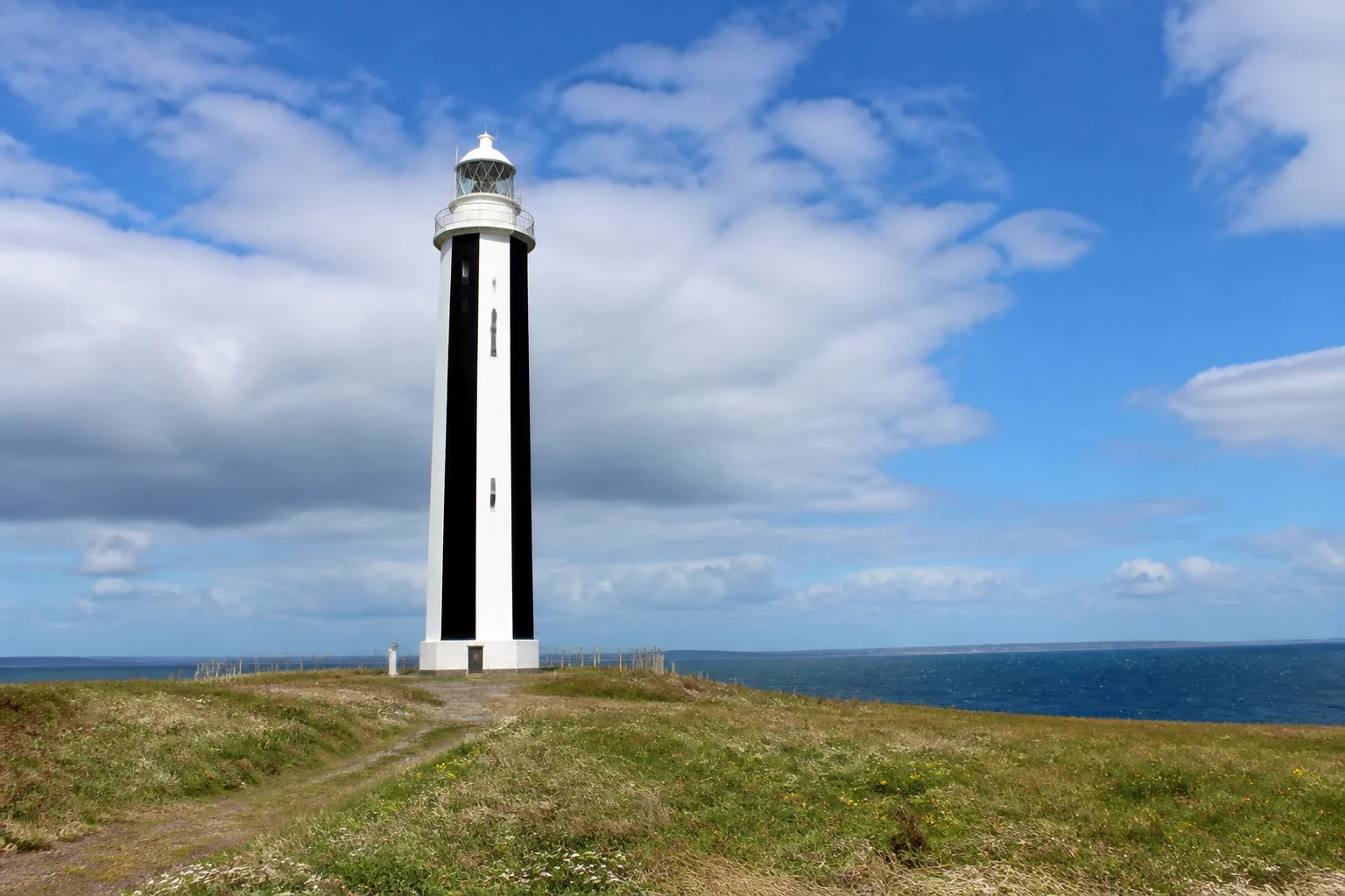 Start Point lighthouse on Sanday, Orkney, with its distinctive bold vertical black-and-white striped tower viewed from the windswept grassy headland under a bright partly-cloudy summer sky