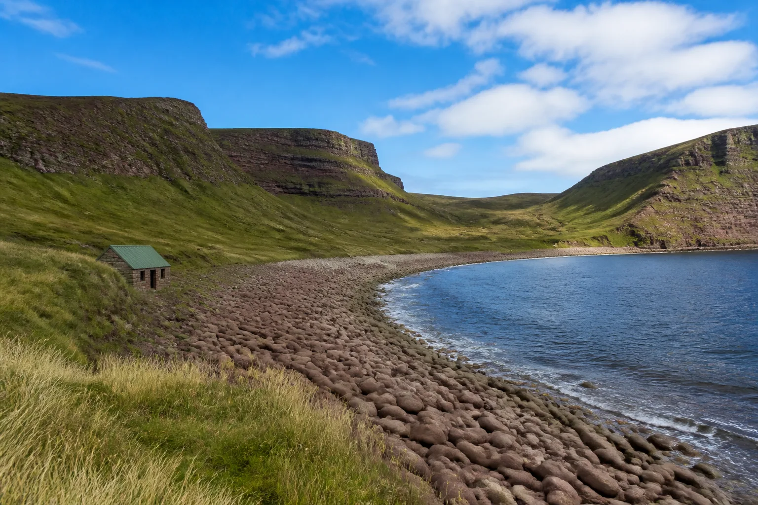 Wide sweeping summer view of Rackwick Bay on the island of Hoy in Orkney, with a curving beach of enormous smooth red-sandstone boulders leading to dark blue sea, steep grass-and-heather cliffs rising on either side, and a single small abandoned stone fisherman's bothy with a green tin roof on the grass above the boulder beach
