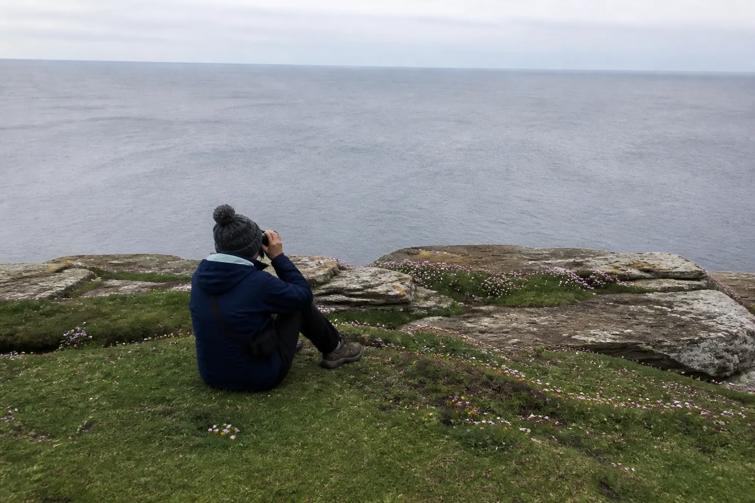 Casual visitor in a navy waterproof jacket and woolly hat sat on the cliff edge at Mull Head, Deerness, Orkney, watching the sea through binoculars at a respectful distance