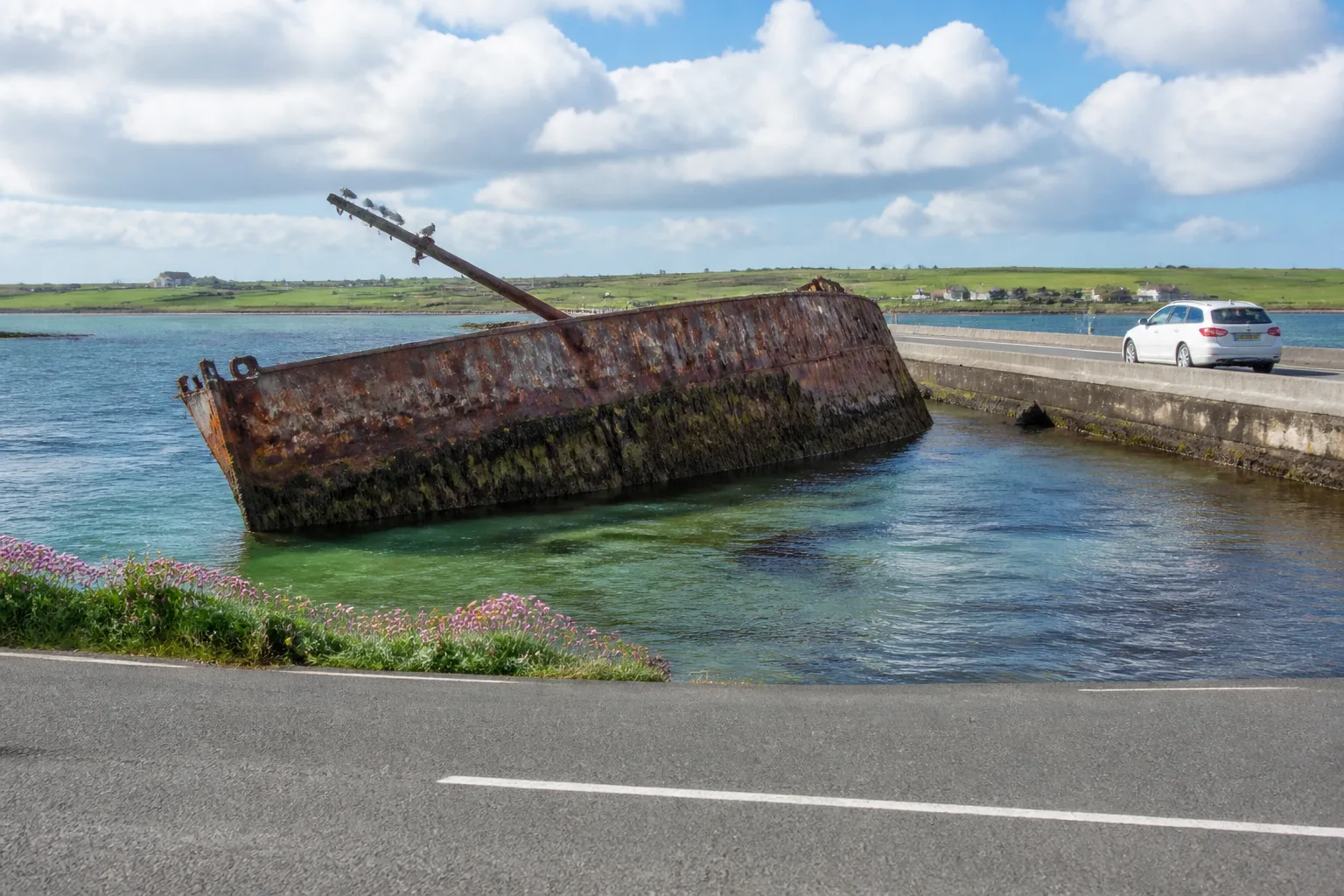 Rusting WWI blockship half-sunk in the turquoise water beside Churchill Barrier No. 3 on Orkney, with the concrete causeway and a single estate car visible on the right and the low green coast of Burray in the distance under bright summer cumulus