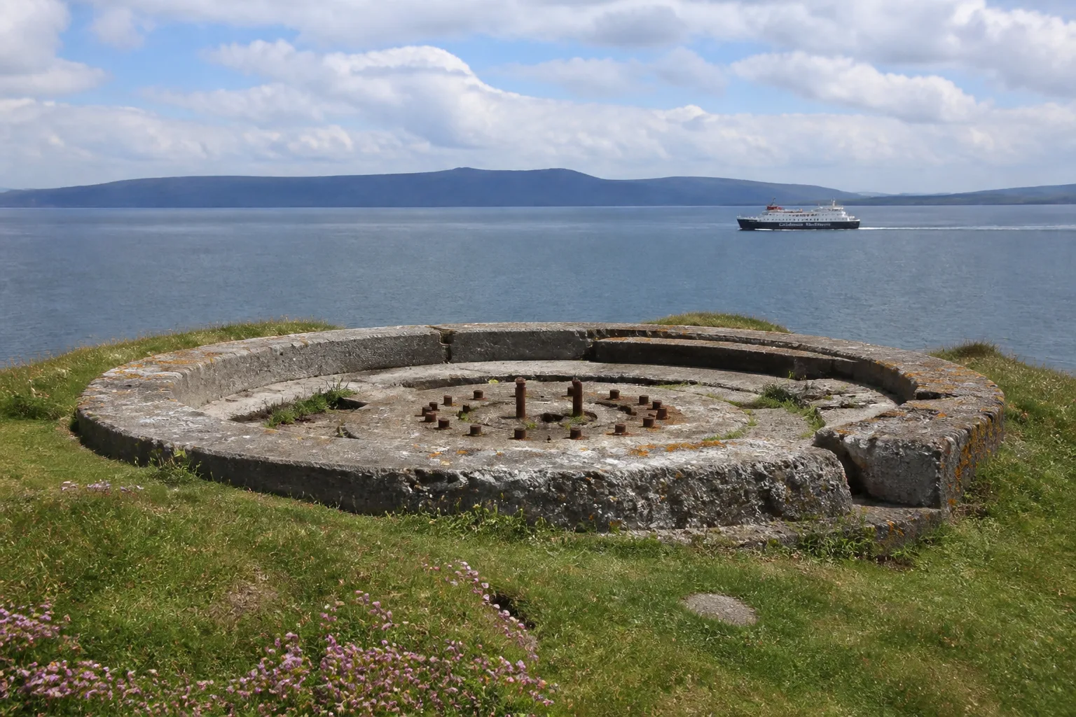 Crumbling concrete WWII coastal gun emplacement at Hoxa Head on South Ronaldsay Orkney — circular gun pit weathered to pale grey with patches of orange lichen, rusting iron mounting bolts in the centre, set in green clifftop turf overlooking the flat steel-blue sea of Hoxa Sound with the dark hills of Hoy on the horizon and a small ferry crossing