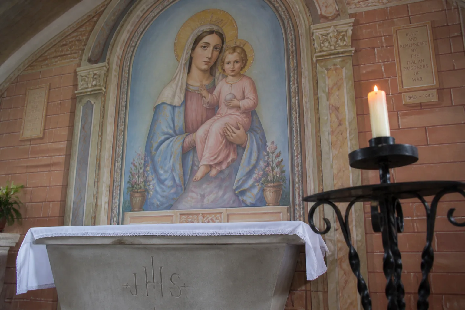 Close-up interior of the Italian Chapel altarpiece on Lamb Holm Orkney — Domenico Chiocchetti's painted fresco of the Madonna and Child in renaissance pinks and blues with a gold halo above the simple concrete altar, with a wrought-iron candle stand and lit votive candle in soft mixed daylight
