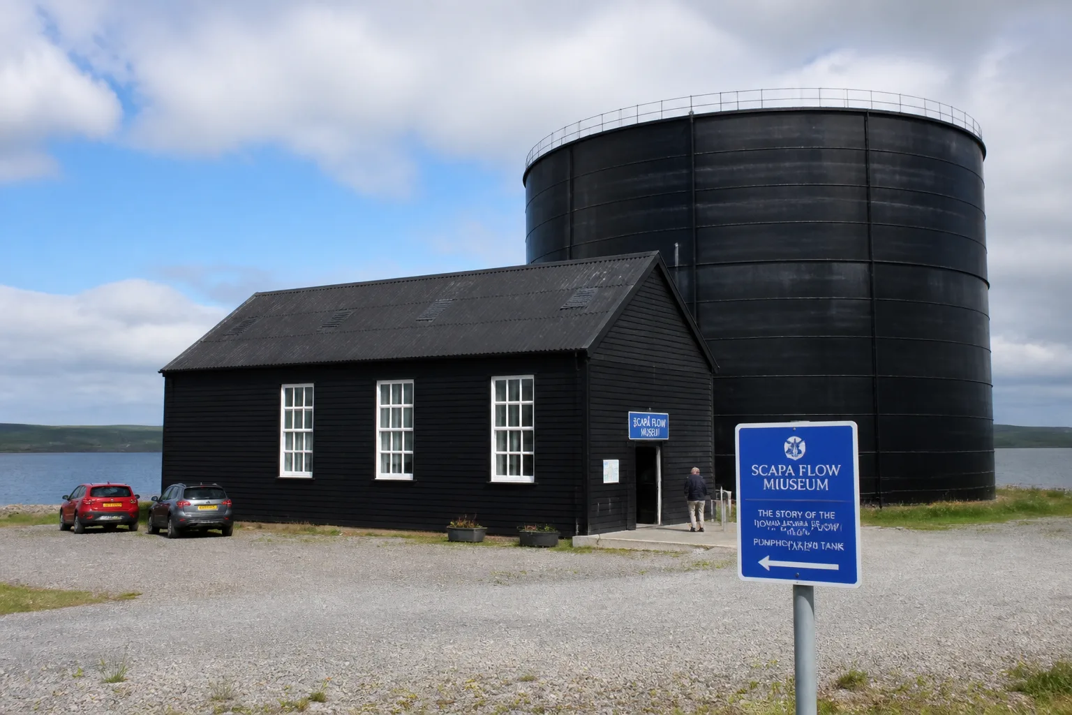 The Scapa Flow Museum at Lyness on the Isle of Hoy Orkney — the black-painted wooden former WWII Royal Navy fuel oil pumphouse with tall white-rimmed windows and pitched corrugated roof beside an enormous black cylindrical oil tank, modern blue museum signage in the foreground, with the calm grey-blue water of Scapa Flow beyond under bright summer cumulus