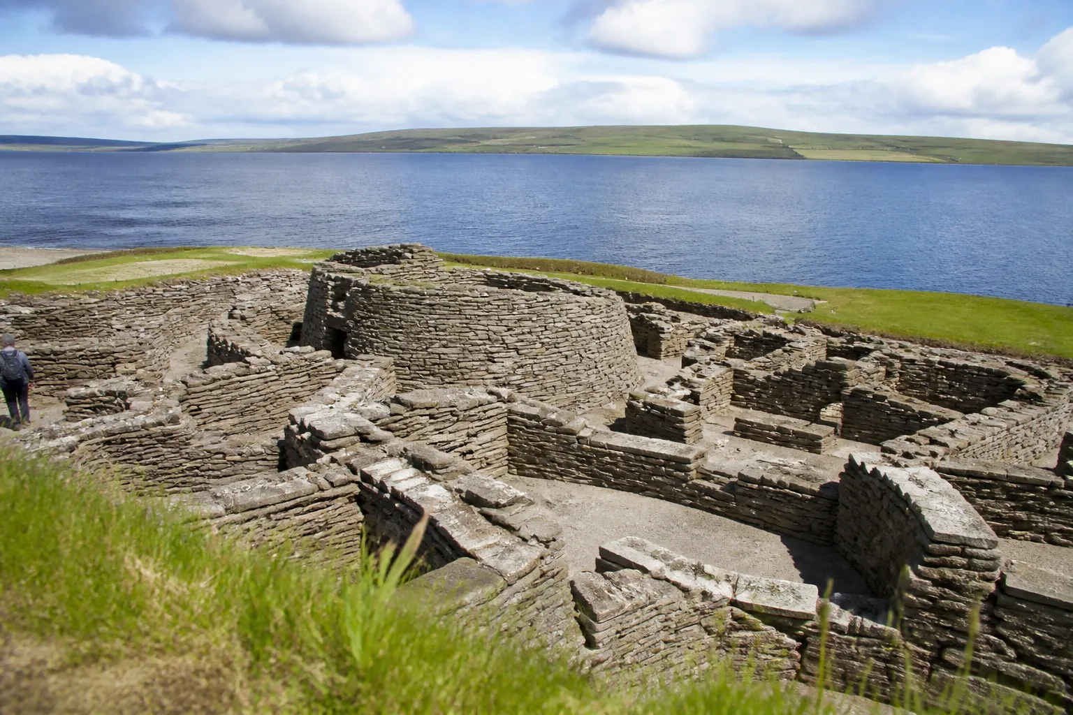 Overhead view of the Pictish-era and Iron Age stone houses around the Broch of Gurness at Evie, Orkney — clustered drystone walls and paved floors arranged around the central broch tower stump, with Eynhallow Sound and the hills of Rousay visible in the background