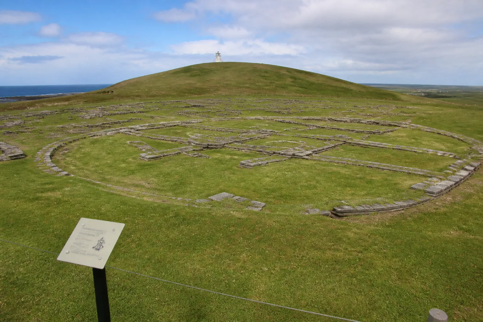 Exposed grass-cropped stone foundations of the Pictish and Norse settlement on the summit of the Brough of Birsay, Orkney, looking up the green tidal island towards the small white modern lighthouse on the highest point