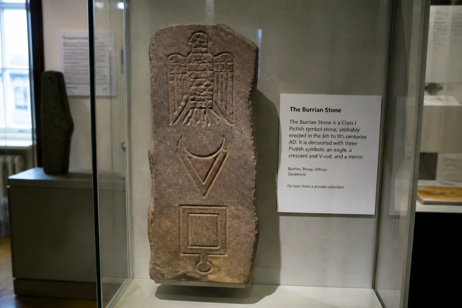 The Burrian symbol stone on display behind glass at the Orkney Museum in Kirkwall — a Class I Pictish slab with carved eagle, crescent and V-rod, and mirror symbols, with a printed museum label beside it