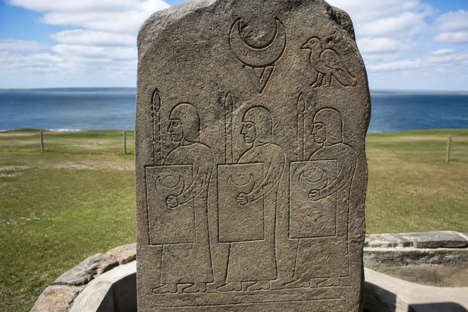 Close-up of the Pictish symbol stone replica on the Brough of Birsay, Orkney — grey-brown sandstone slab carved with three robed Pictish warrior figures carrying square shields and spears, with a crescent and V-rod symbol and an eagle visible above