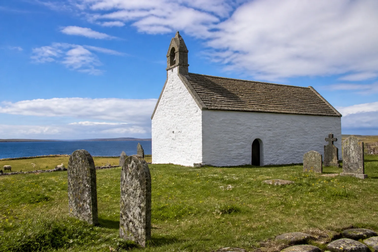 The white-harled medieval St Boniface Kirk on Papa Westray, Orkney — a small rectangular stone kirk standing alone in a sloping clifftop graveyard with weathered grave-slabs in the foreground and the North Atlantic horizon visible behind