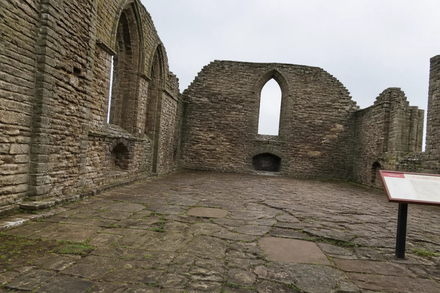 Roofless ruins of the Earl's Palace in Kirkwall, Orkney — empty stone window arches and crumbling sandstone walls open to a flat grey sky, with a flagstone floor weathered by centuries of exposure