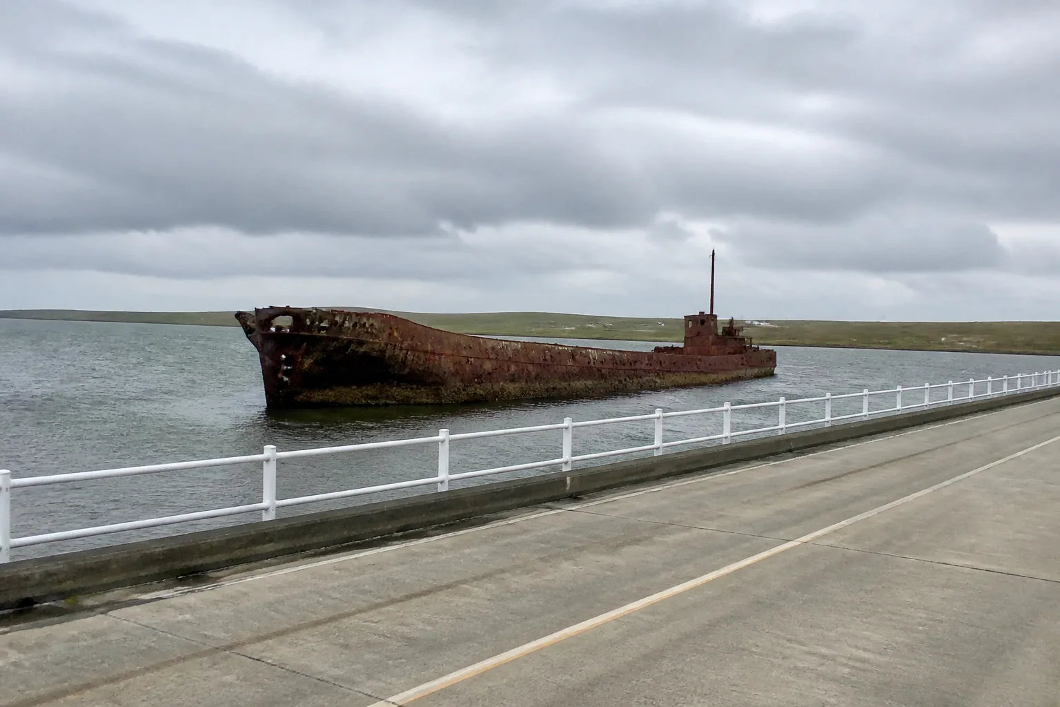 Rusting WWI blockship half-sunk in the channel beside Churchill Barrier No. 2, Orkney — reddish-brown rust-streaked steel hull lying at an angle in the grey-green water with kelp clinging to its sides
