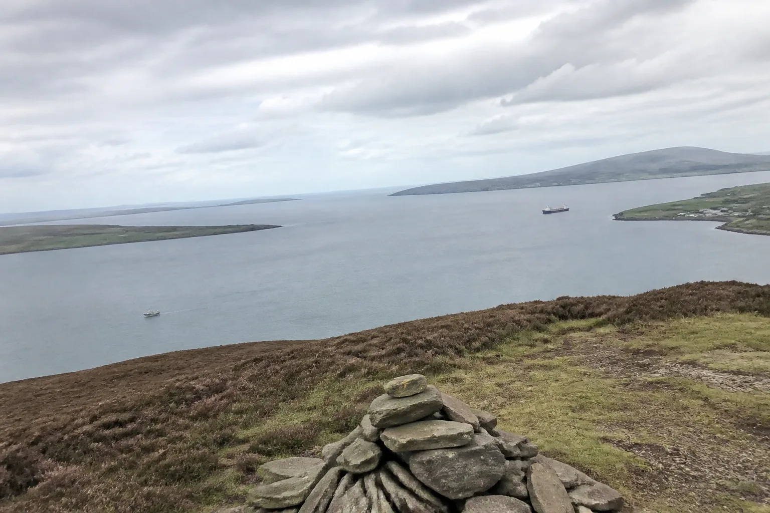 Scapa Flow seen from the heather-covered top of Wideford Hill above Kirkwall — the vast natural harbour ringed by Hoy and South Ronaldsay, with a small stone cairn in the foreground and a distant tanker on the calm grey water