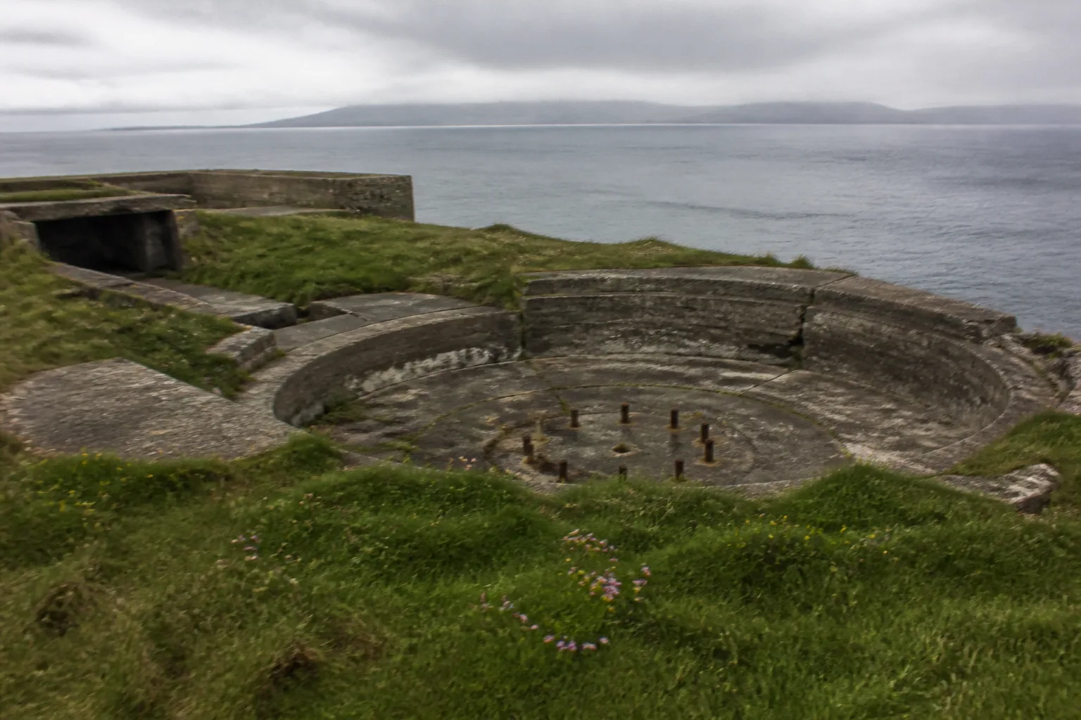 Crumbling concrete WWII coastal gun emplacements at Hoxa Head, South Ronaldsay, Orkney — circular gun pits set into the green clifftop turf overlooking the entrance to Scapa Flow with rusting iron mounting bolts where the artillery once sat
