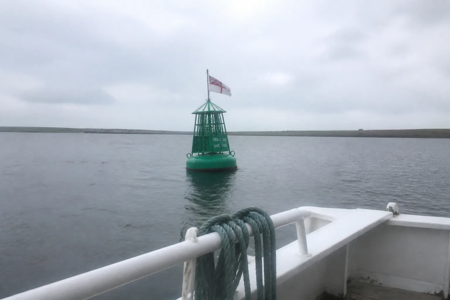 The green navigational buoy floating on the calm grey water of Scapa Flow that marks the war grave of HMS Royal Oak, with a small Royal Navy ensign attached, photographed from the deck of a small fishing boat