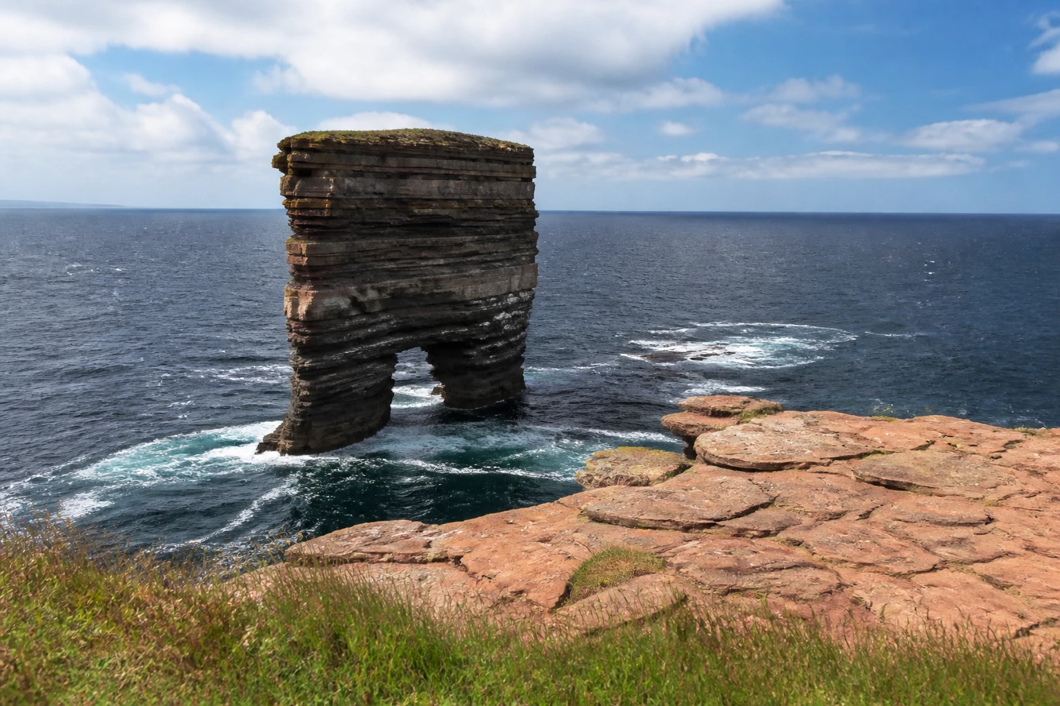The two-legged Yesnaby Castle sea stack on the west coast of Orkney Mainland, a wide column of layered Old Red Sandstone with a clear arch through the lower portion forming two leg-like supports, photographed from the layered red sandstone clifftop with the choppy Atlantic in the background under a bright partly-cloudy summer sky