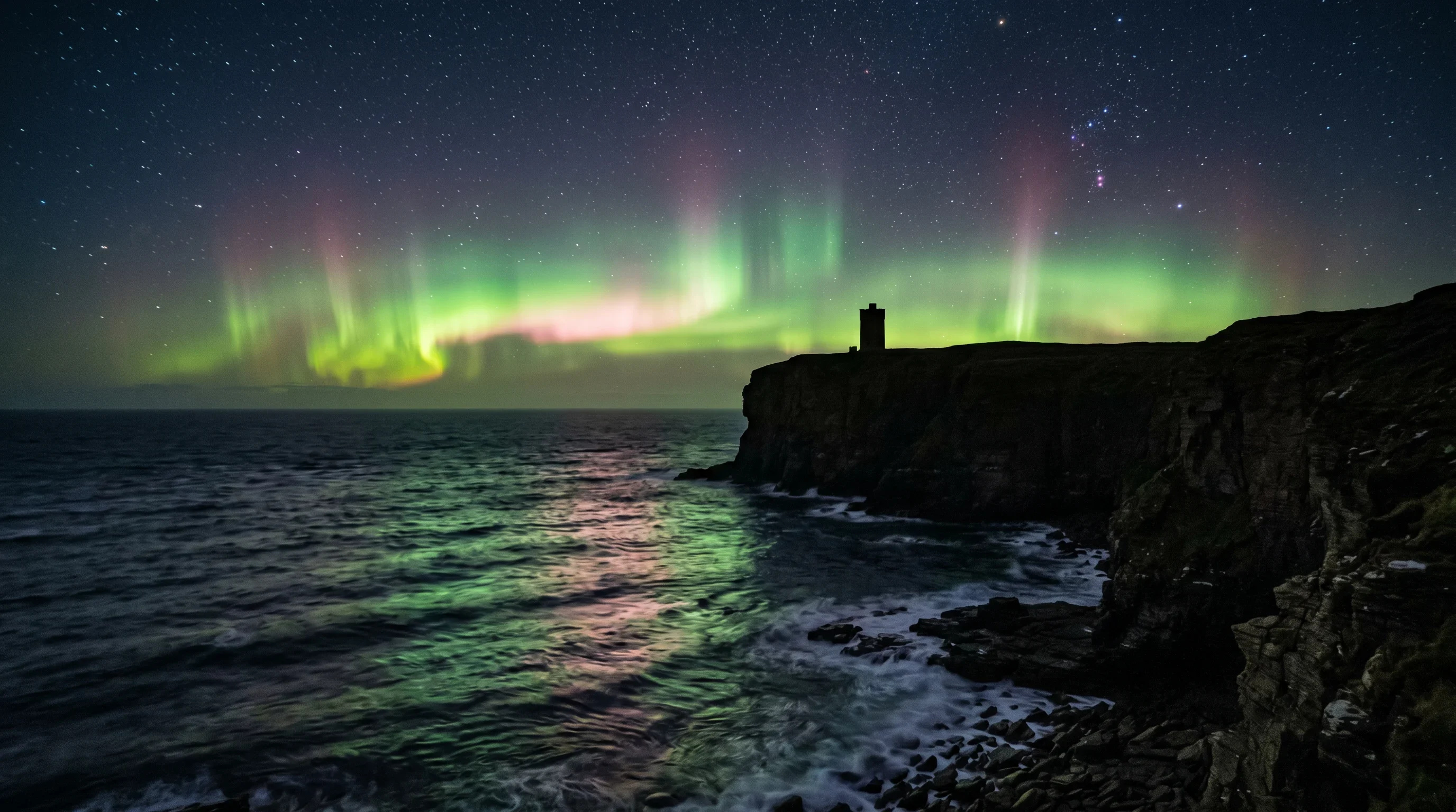 The Aurora Borealis reflecting off the dark Atlantic below the cliffs at Marwick Head, Orkney, with the Kitchener Memorial tower silhouetted against the green and pink light