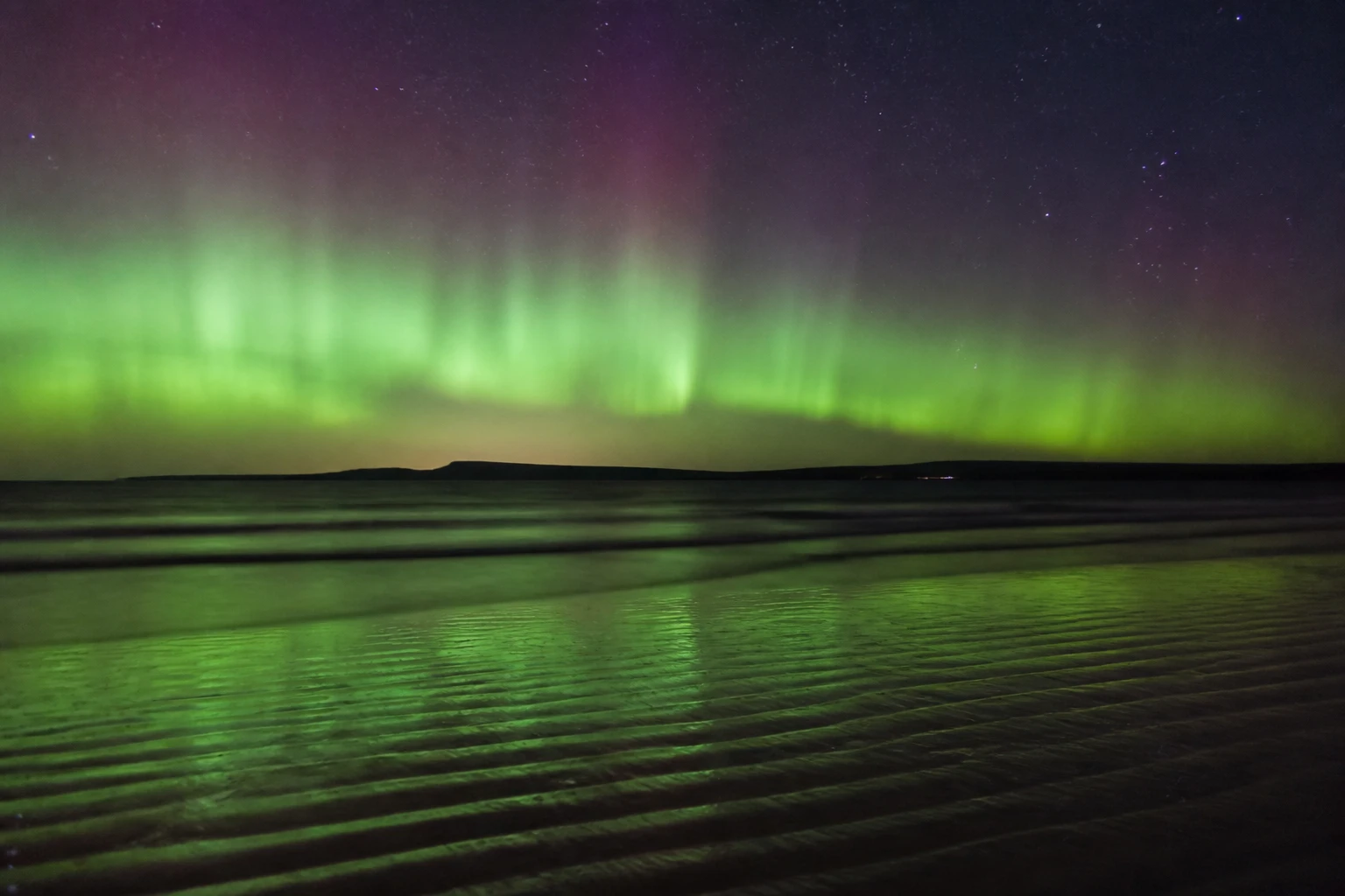 Long-exposure night photograph at Sands of Evie on Orkney's north coast with a strong Northern Lights aurora display of green vertical curtains rising from the horizon and a soft pink-magenta wash at the upper edge, the green light reflected on damp rippled sand in the foreground, the silhouette of the Brough of Birsay or Rousay across the dark sea on the horizon under a starry deep navy sky