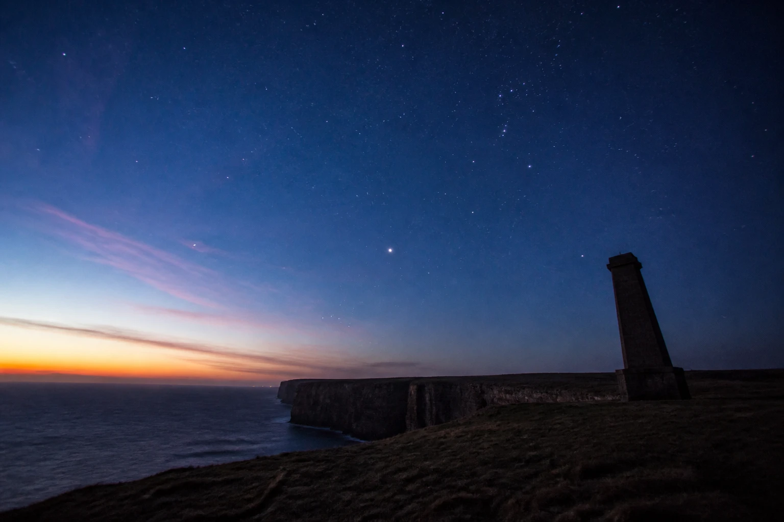 Long-exposure photograph at Marwick Head on the west coast of Orkney Mainland during nautical twilight on a clear winter evening showing the dark silhouette of the Kitchener Memorial tower on the cliff edge against a deepening blue gradient sky with the first dozen brightest stars including Orion's belt low in the south-east emerging, dramatic vertical sandstone cliffs descending to the dark sea, faint pink residual sunset light at the western horizon