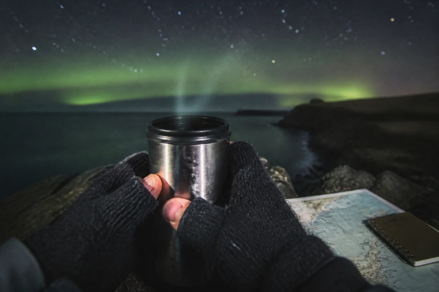 Close-up zoom photograph at night on an Orkney coastal cliff showing two gloved hands in dark thick mittens with cut-off fingertips cradling a battered stainless-steel travel thermos with visible steam rising into the cold air, a faint green band of Northern Lights aurora and scattered stars visible in soft focus in the background, a folded map and small notebook resting on a rock beside the hands