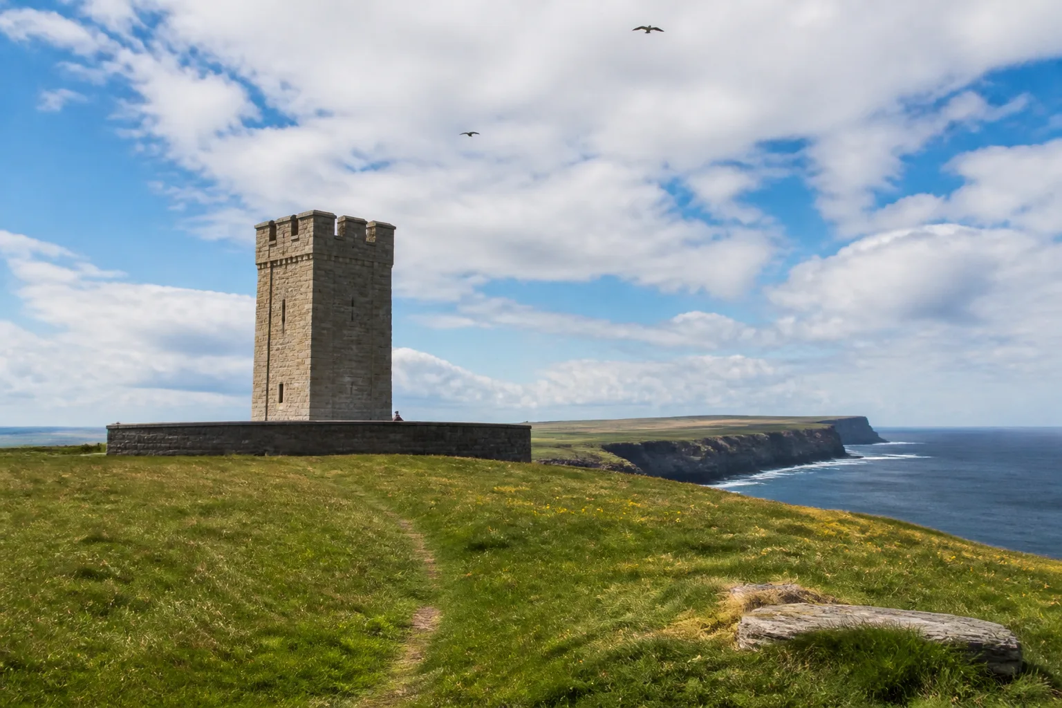Mid-range photo of Marwick Head on West Mainland Orkney looking across green clifftop sheep-grazed pasture towards the Kitchener Memorial — a square crenellated stone tower roughly 15 metres tall built of pale Orkney sandstone standing alone on the cliff edge with a low semicircular memorial wall of darker granite around it, the 87-metre cliffs dropping away to the sea behind under a bright partly-cloudy June afternoon sky