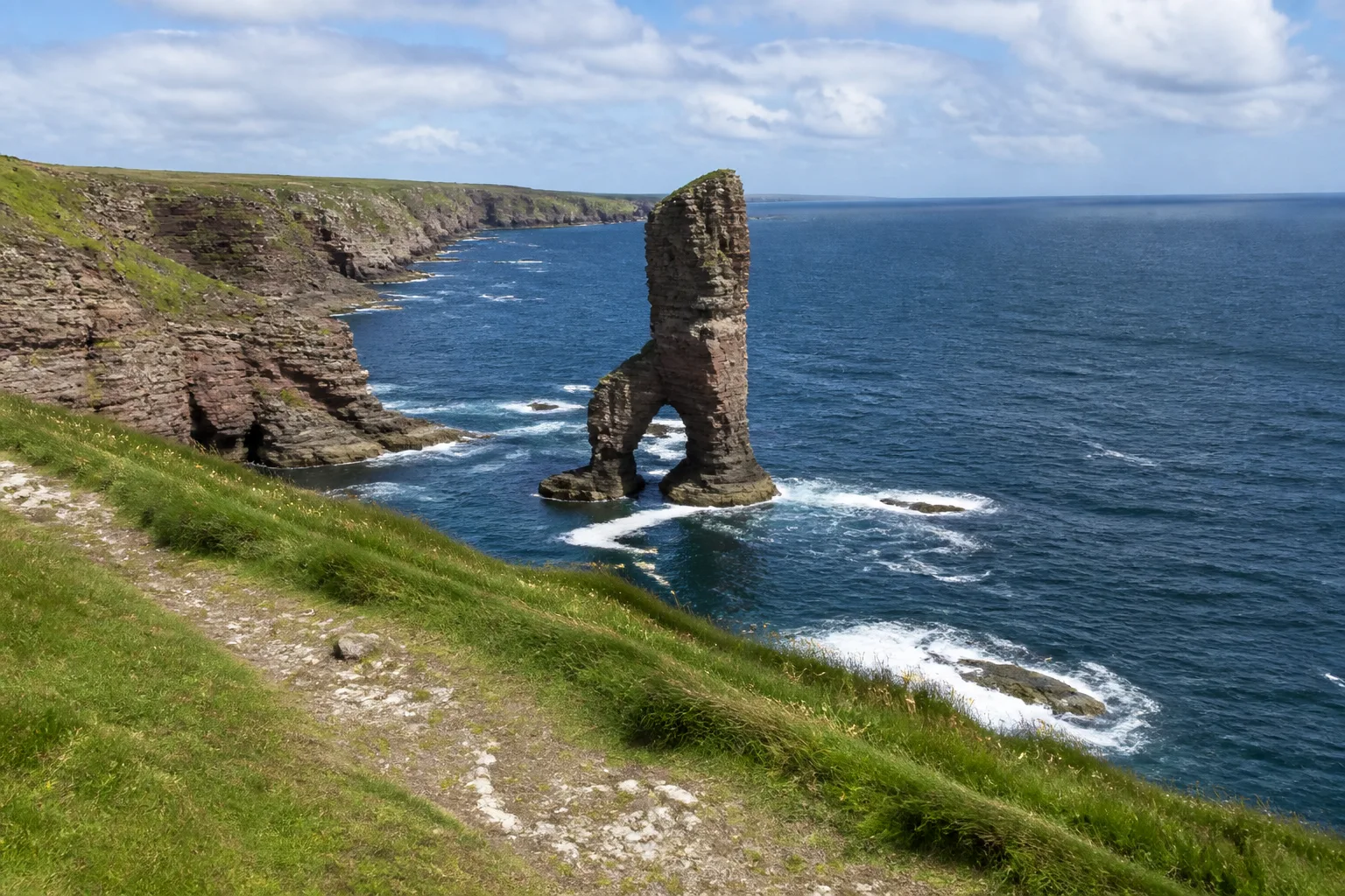 Mid-range photo from the cliff path at Yesnaby on Mainland Orkney looking south along the dramatic Old Red Sandstone coastline with the Castle of Yesnaby sea stack in the centre of the frame — a delicate narrow sea stack about 35 metres high with a natural arch at its base resting on a split rock pedestal just offshore from the layered orange-pink and grey sandstone cliffs, with a narrow grassy walking path in the foreground under a partly-cloudy June day