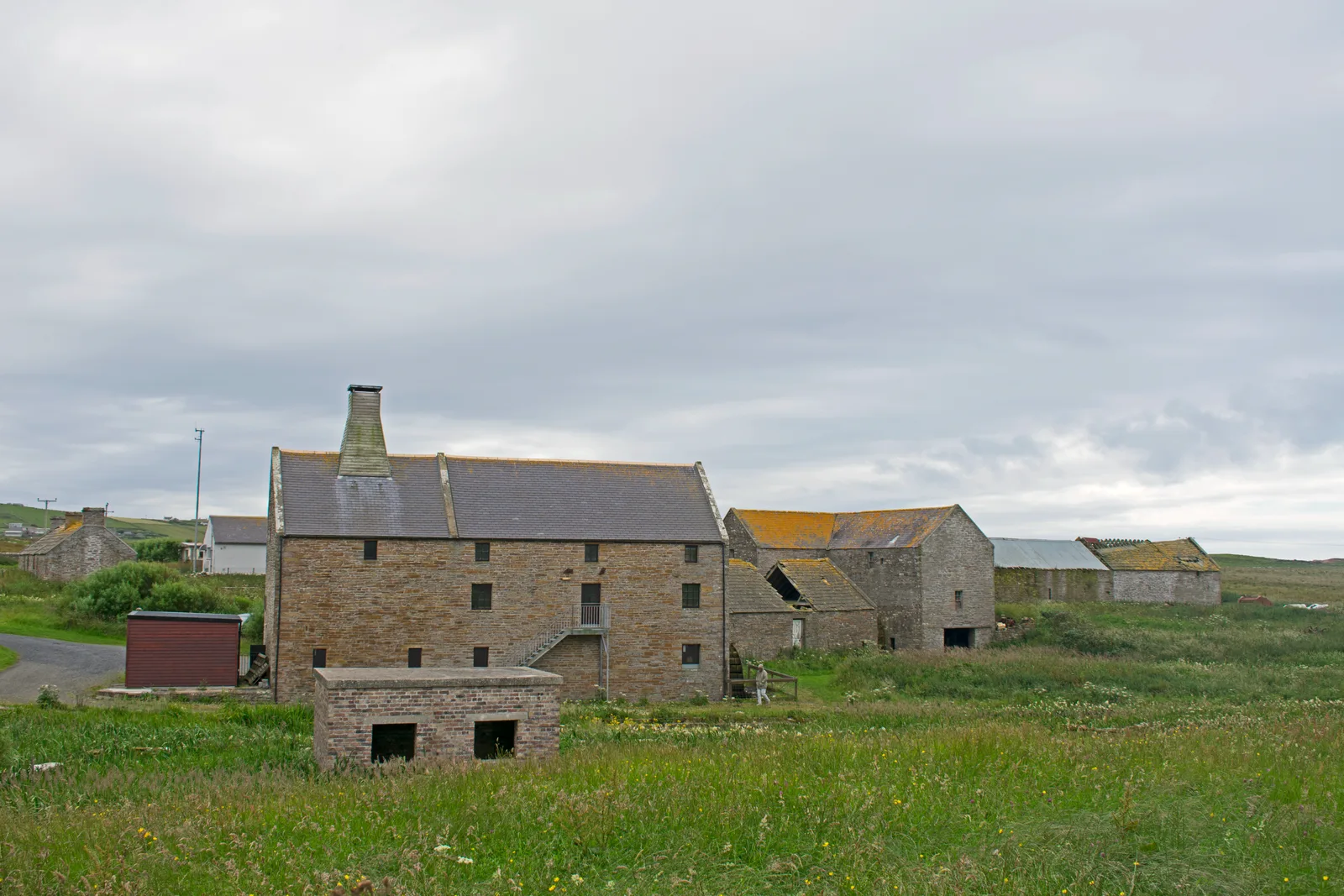Barony Mill at Birsay seen from the north across rough grass — a three-storey flagstone mill building with a tapered kiln vent on the roof ridge and an external stair, with stone outbuildings and steadings running away behind it
