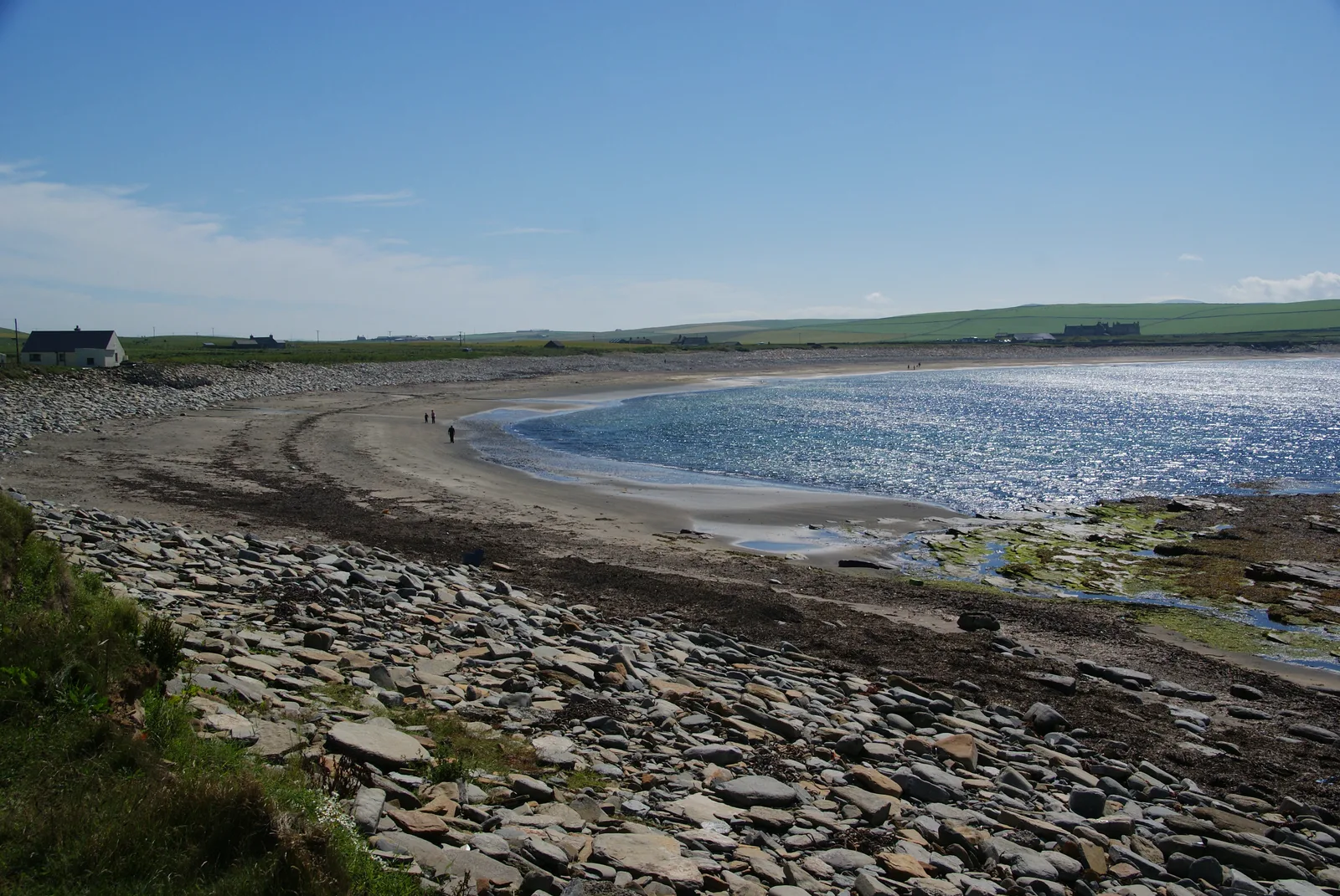 The Bay of Skaill in bright sunshine — a wide curving arc of pale sand with sunlight glittering on the water, a boulder storm beach in the foreground, a white cottage on the low shore at the left and green farmland running back from the bay