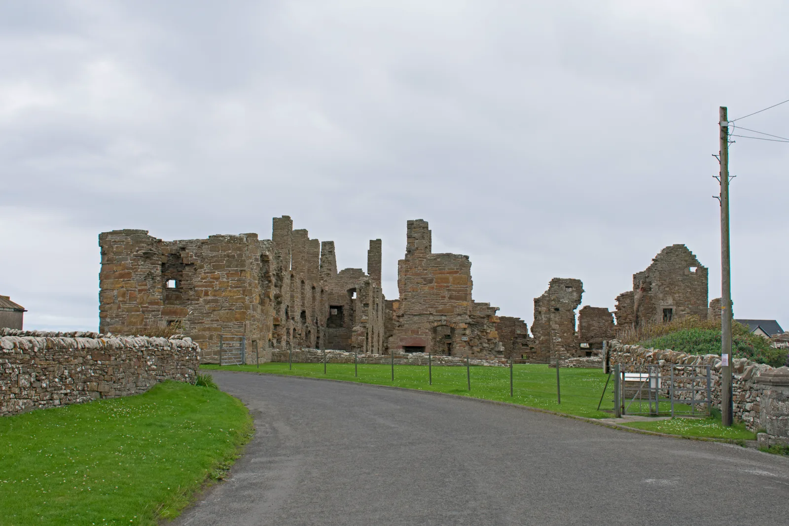 The roofless ruin of the Earl's Palace at Birsay seen from the road, its sandstone walls standing to full height with empty window and doorway openings, behind a drystone dyke under a grey sky